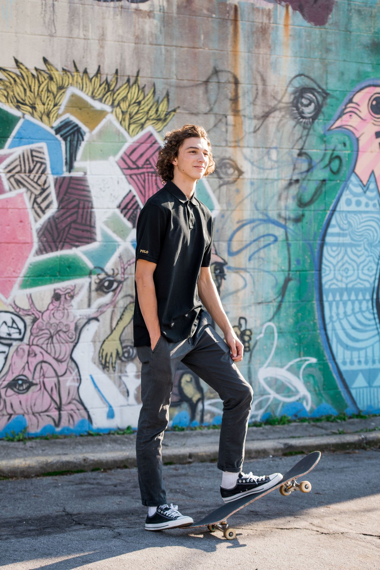 Skateboarder in black attire poses against colorful graffiti wall with urban street art designs.