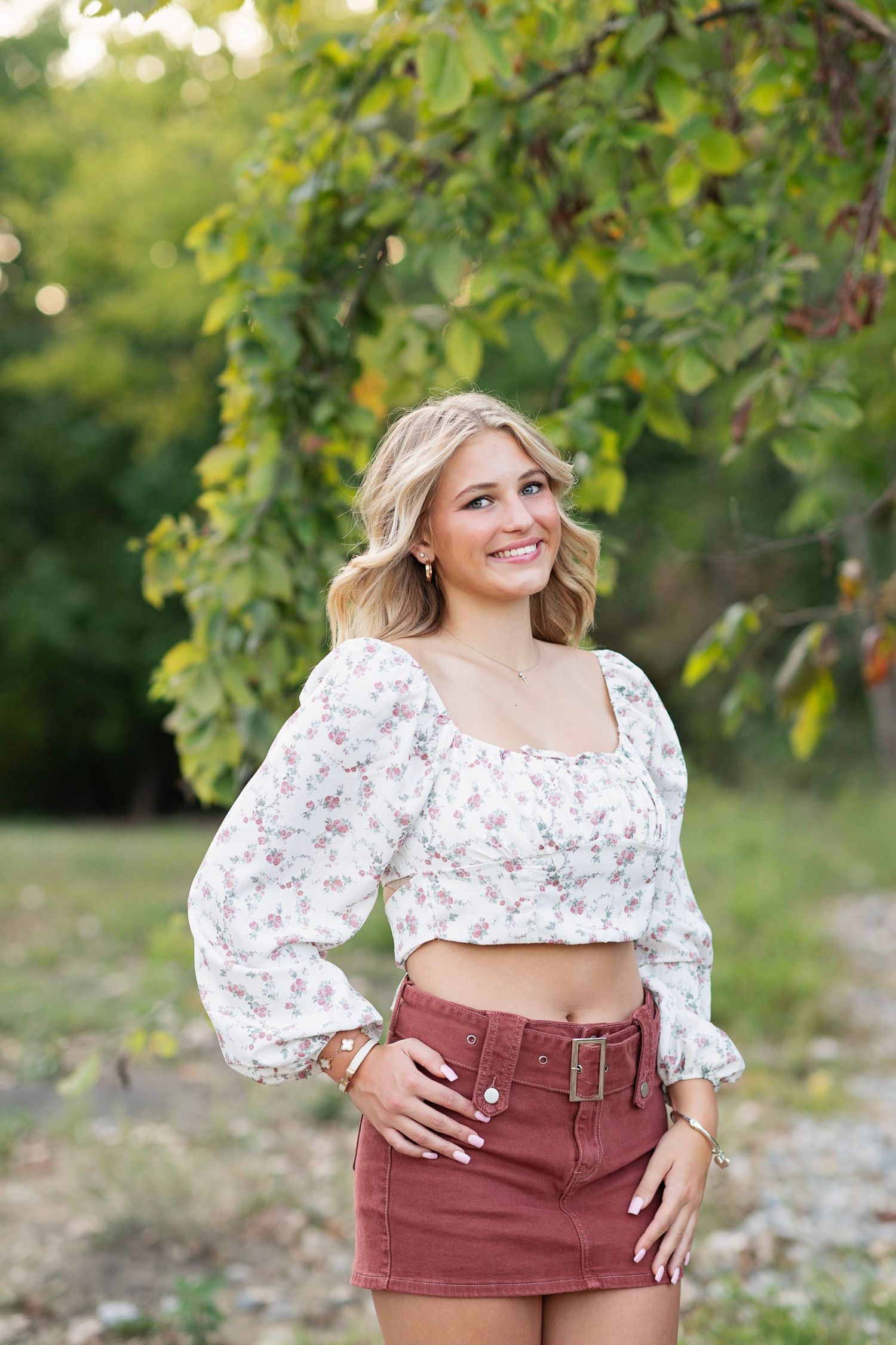 Woman in floral crop top and burgundy skirt poses outdoors under green trees in natural sunlight.