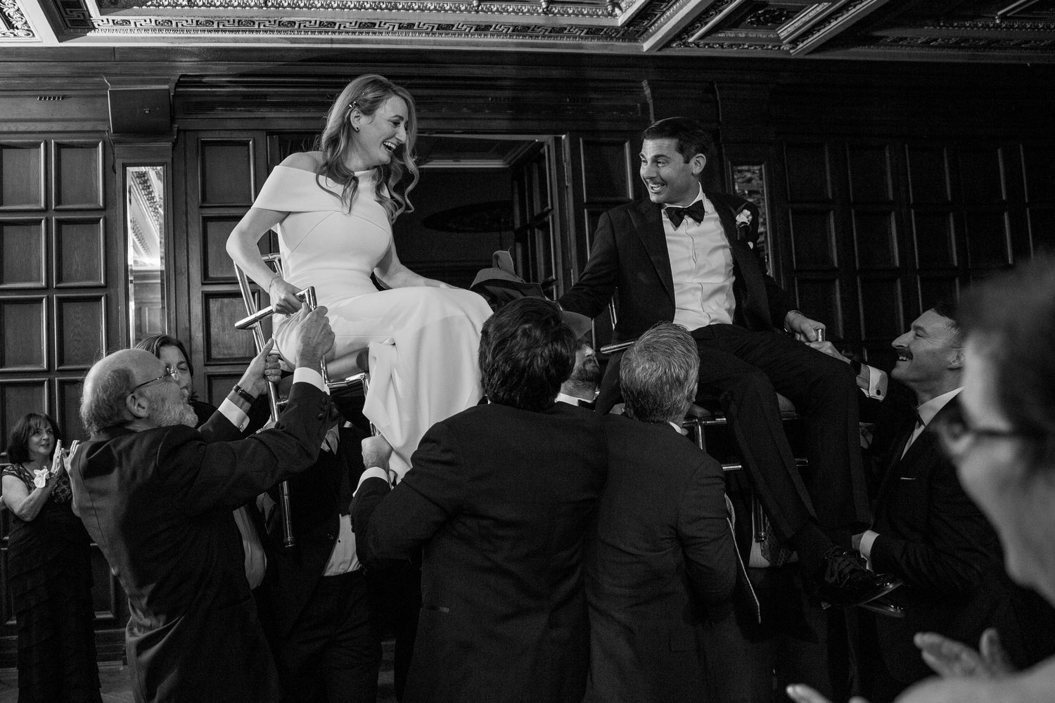 Black and white photo of wedding celebration with guests lifting couple on chairs.