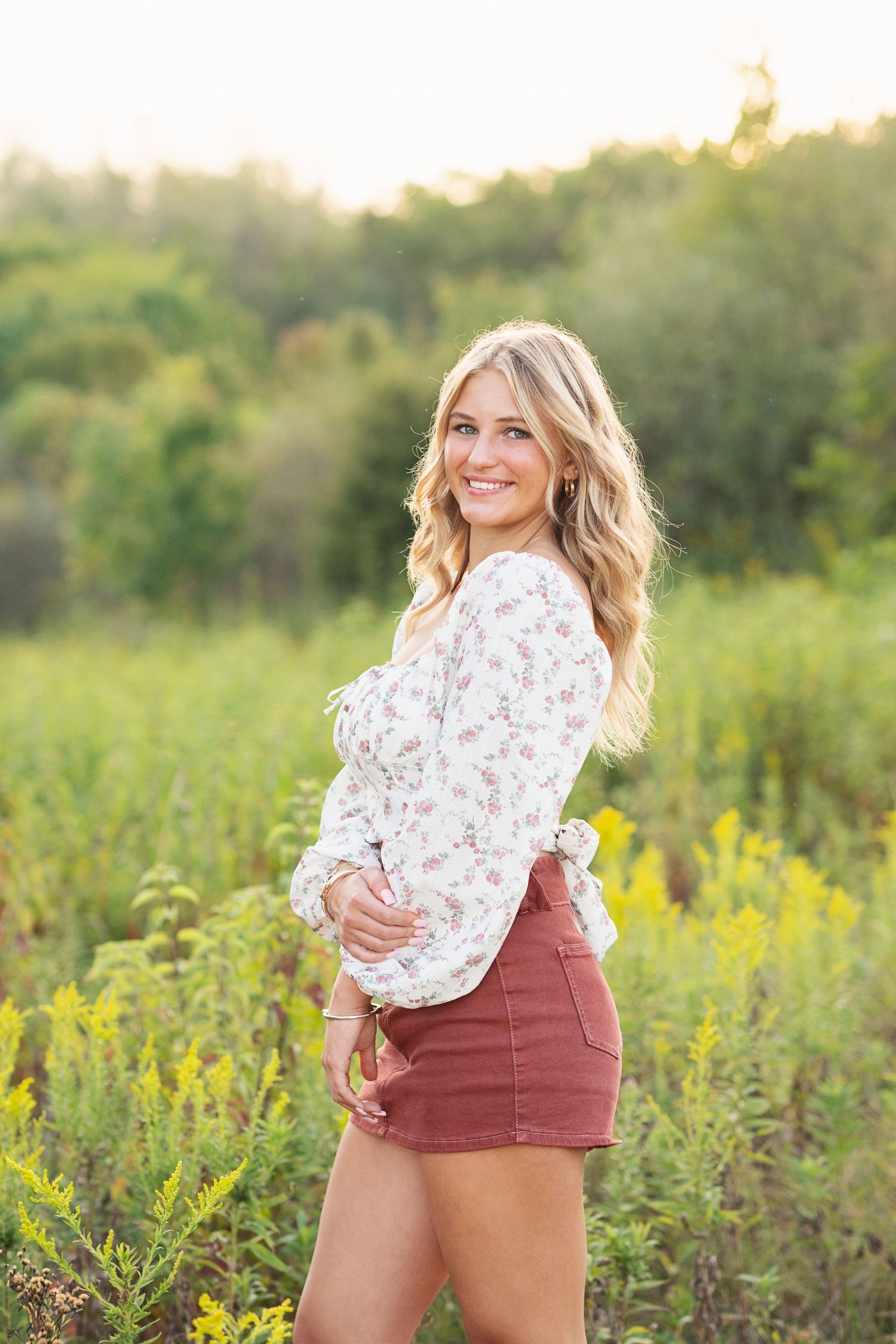 A senior in denim shorts and floral top poses in a sunlit meadow with yellow wildflowers at sunset. Senior session, Galena, OH, Big Walnut.