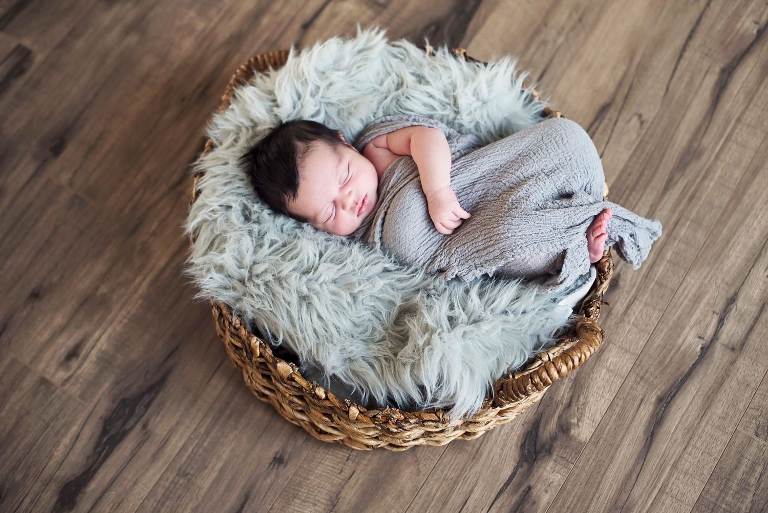 A sleeping newborn wrapped in gray swaddle rests peacefully in a woven basket with fluffy gray fur lining on hardwood floor.