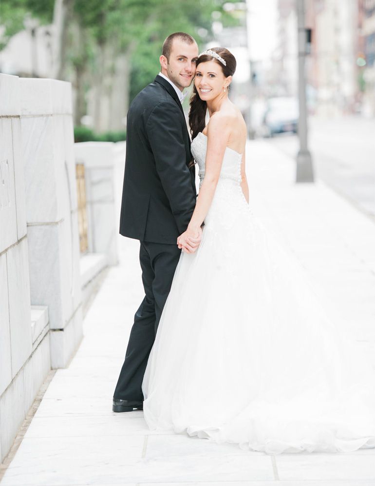 A couple in wedding attire share intimate moments along a white stone wall in an urban setting.