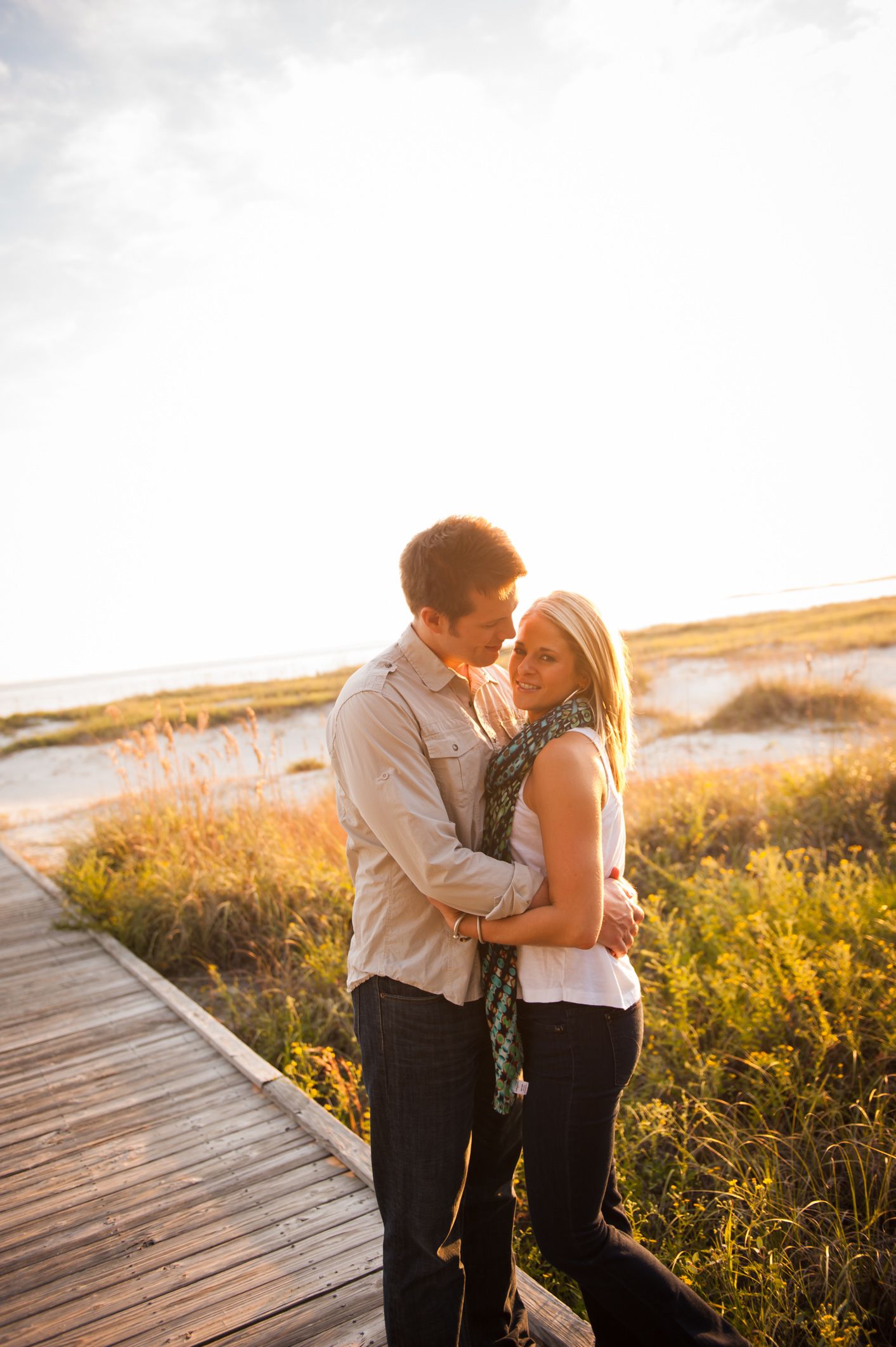 A romantic couple embraces on a wooden boardwalk during golden hour with wildflowers and sunlight in the background.