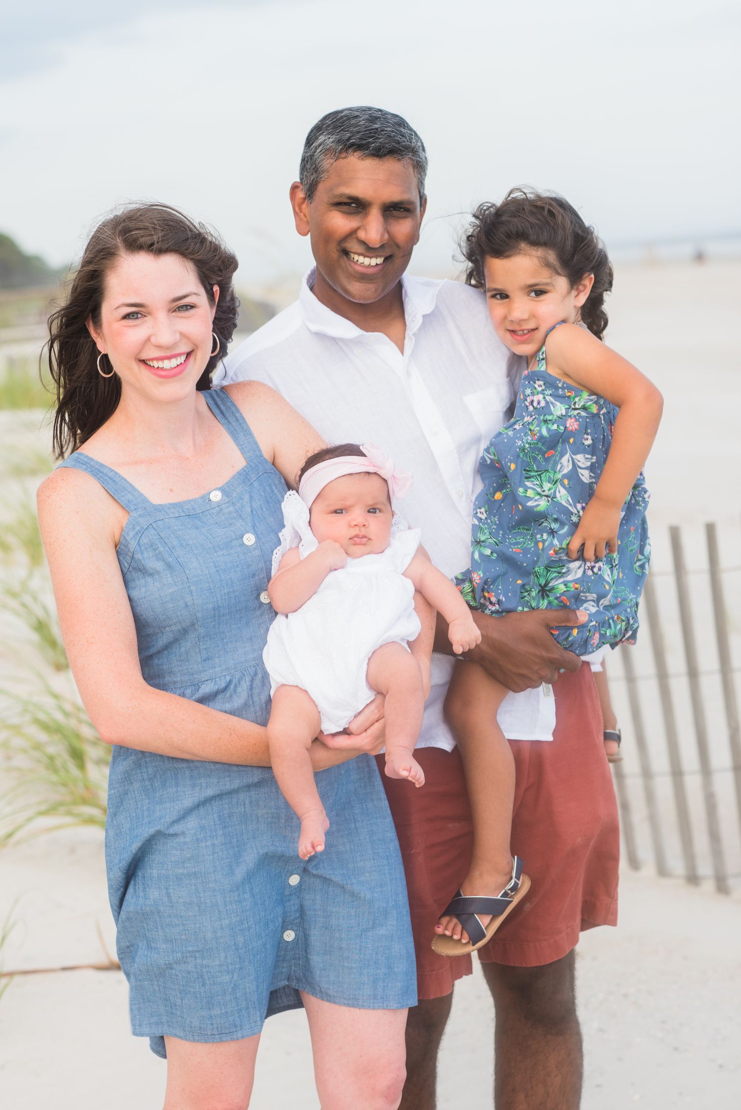 Happy interracial family of four posing on a sandy beach, parents holding two young children on a cloudy day.
