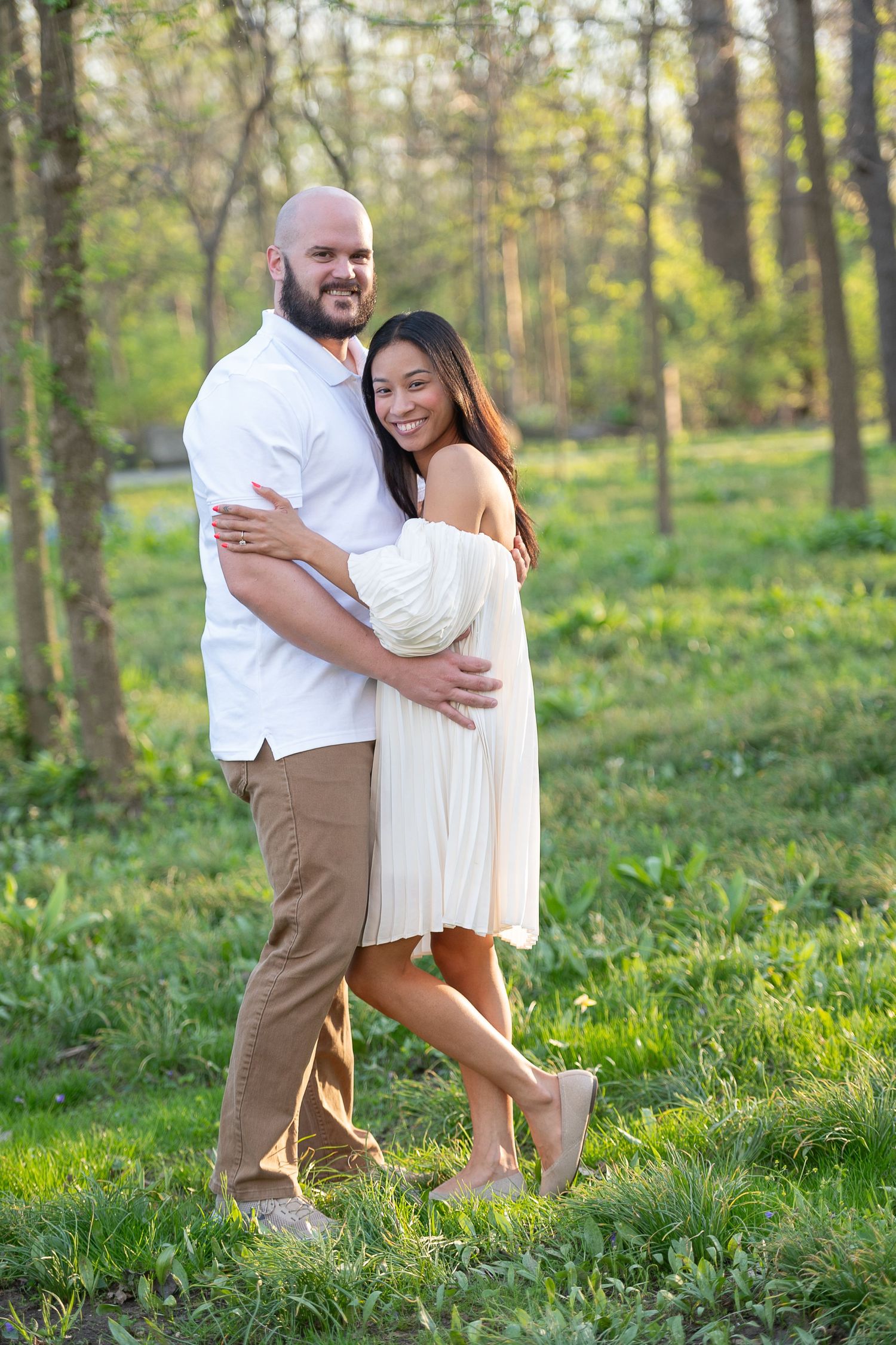 Romantic couple embraces playfully in a sunlit forest setting wearing coordinated white and tan outfits.