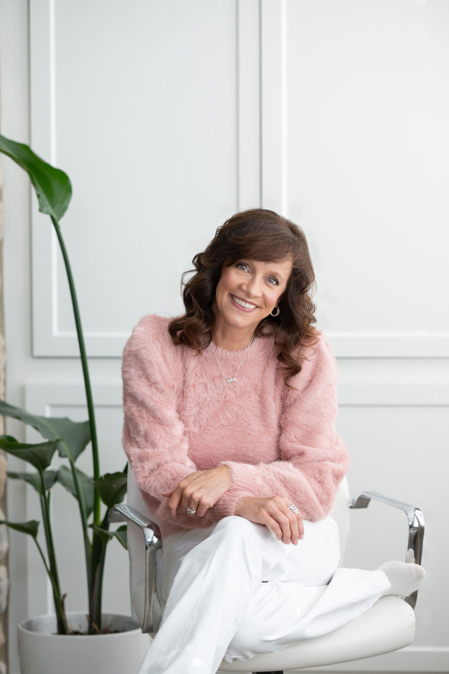 Person wearing a cozy pink sweater relaxes in a bright white room with green plants in the background.