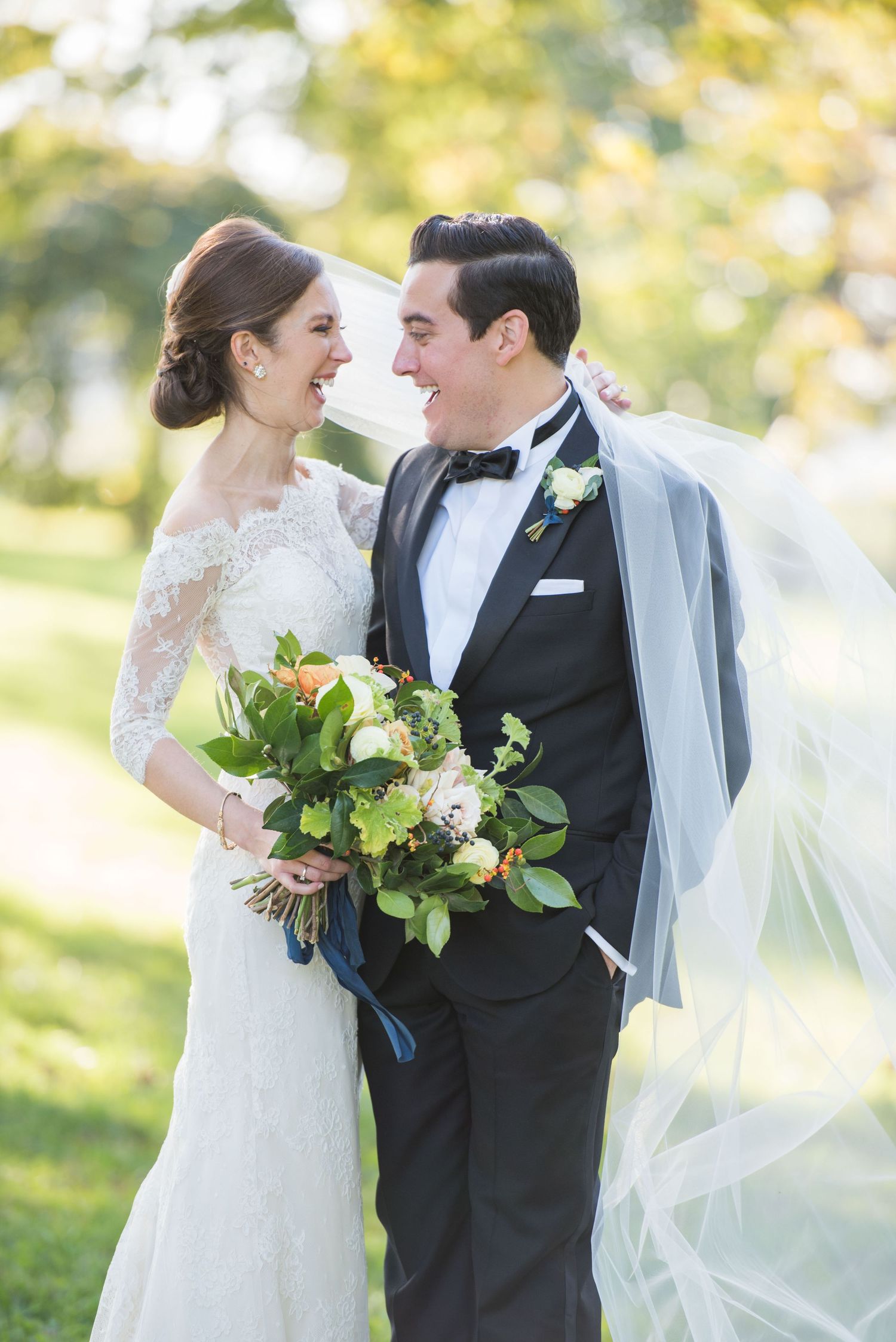 Bride and groom laughing together outdoors, she holds a lush bouquet, veil flowing, in lace gown and black tuxedo.