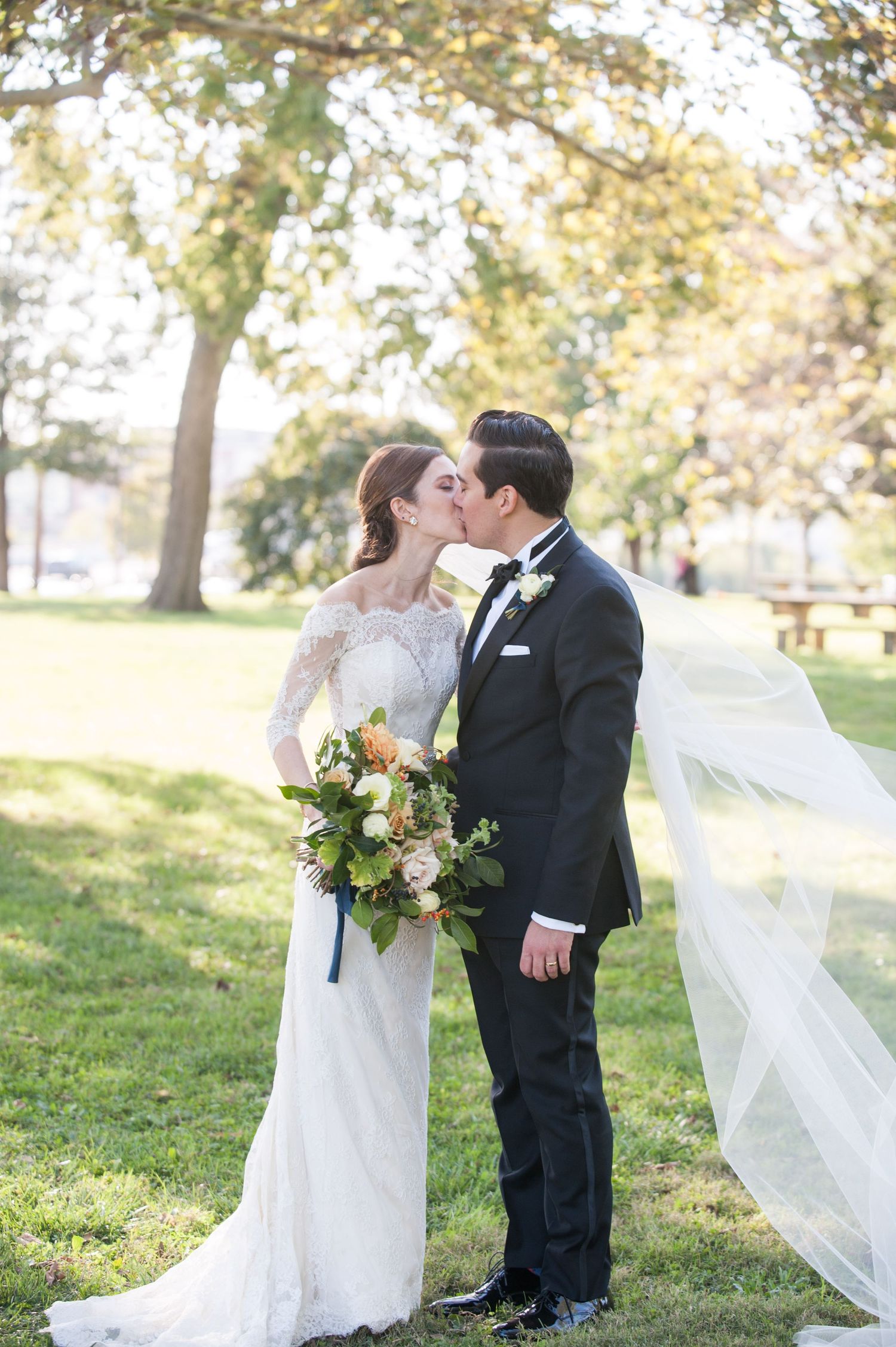 Bride and groom share a kiss outdoors, bride in lace gown with bouquet, groom in black tuxedo, veil flowing in breeze.