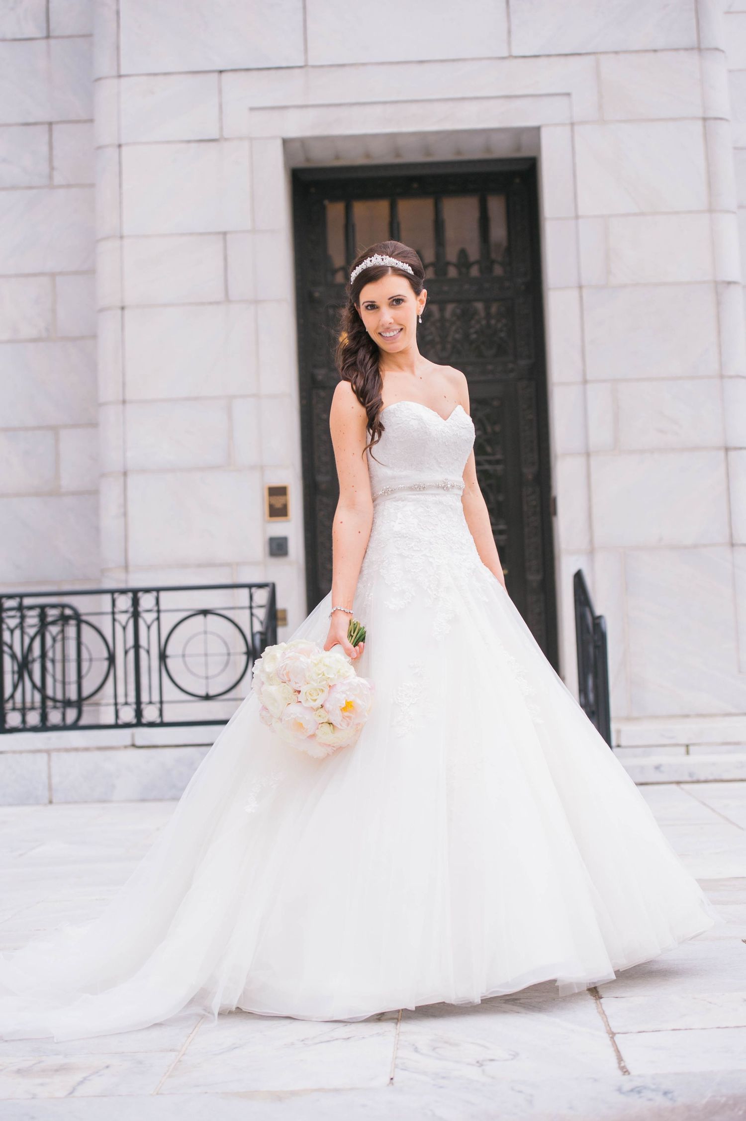 A bride wearing a classic white wedding gown poses against a white stone building exterior.
