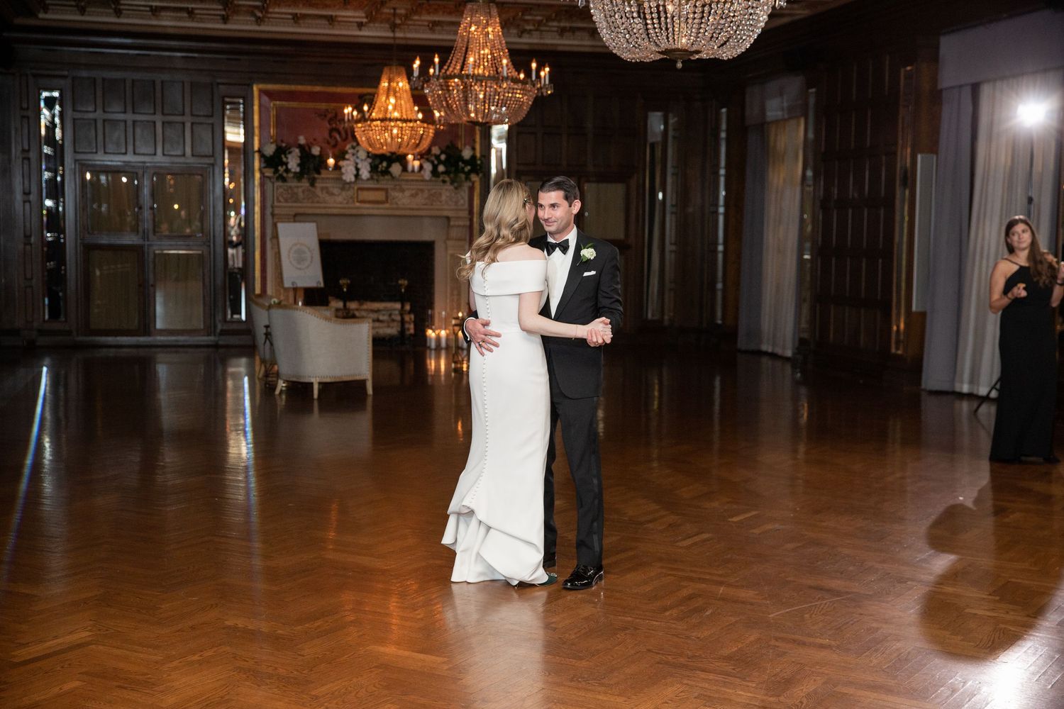 Couple sharing their first dance in an ornate ballroom with wooden floors and chandeliers.