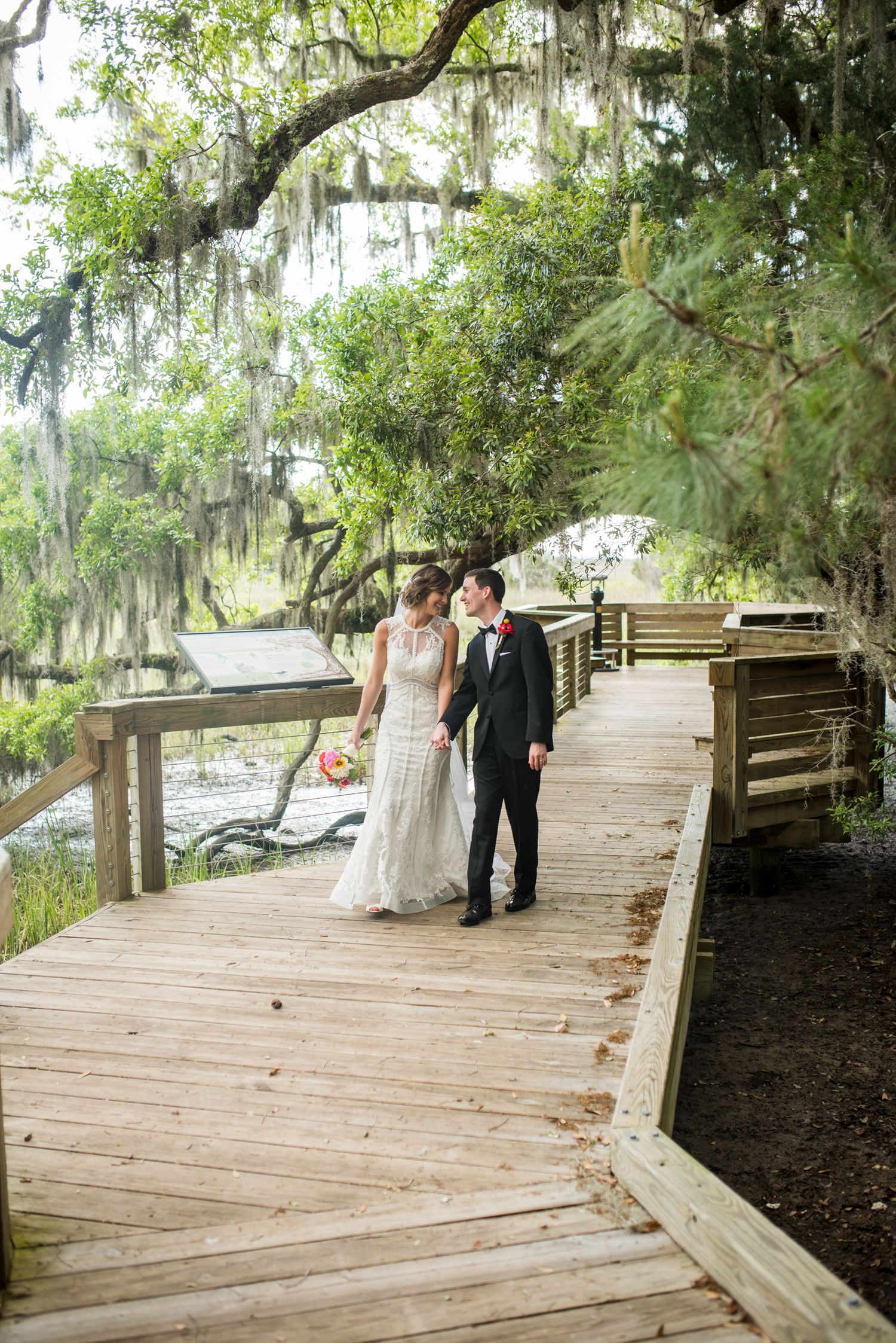 A wooden boardwalk path winds through moss-draped oak trees in a Southern garden setting.