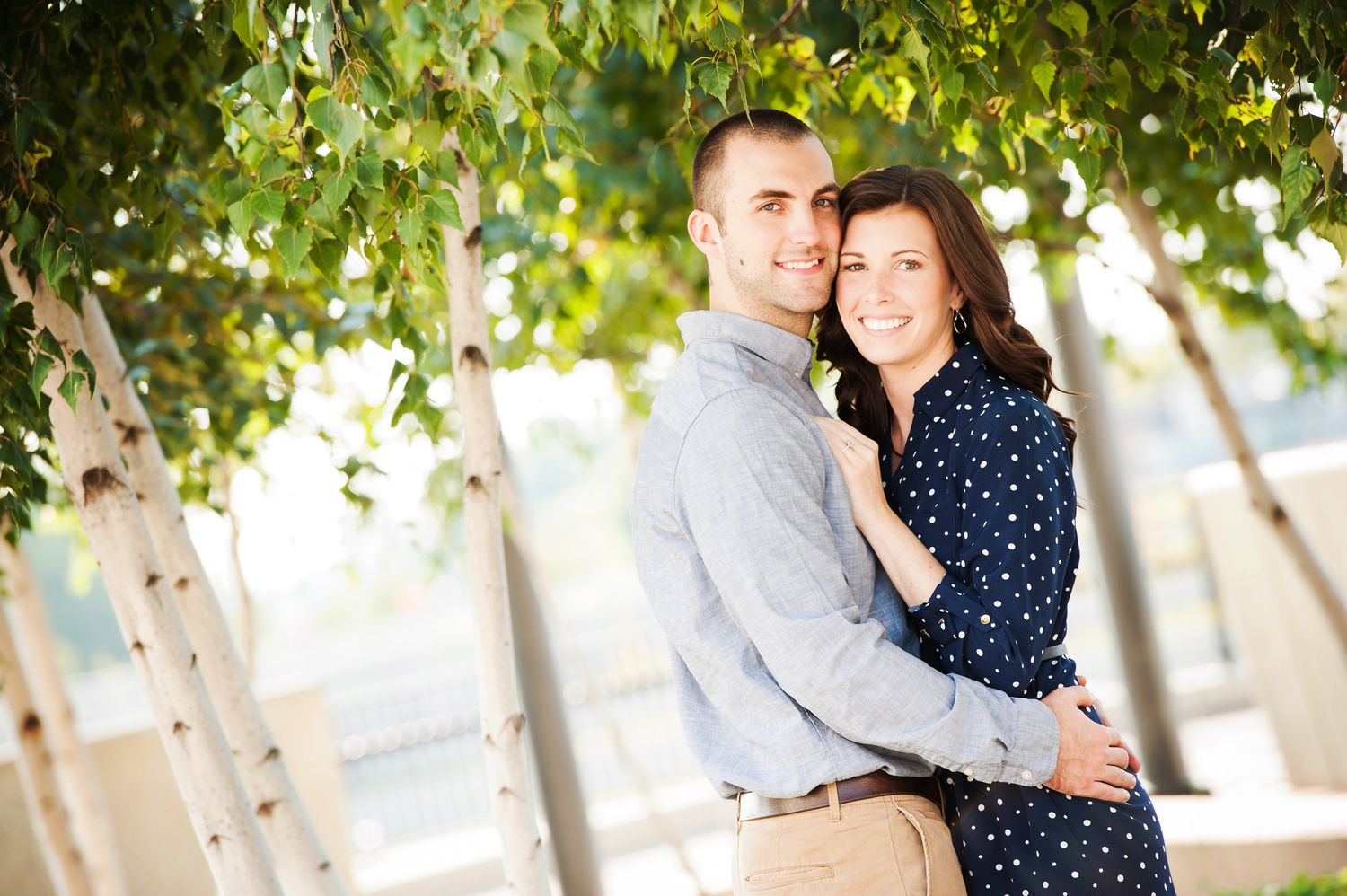A couple embraces under green leaves on a sunny day while wearing business casual attire.