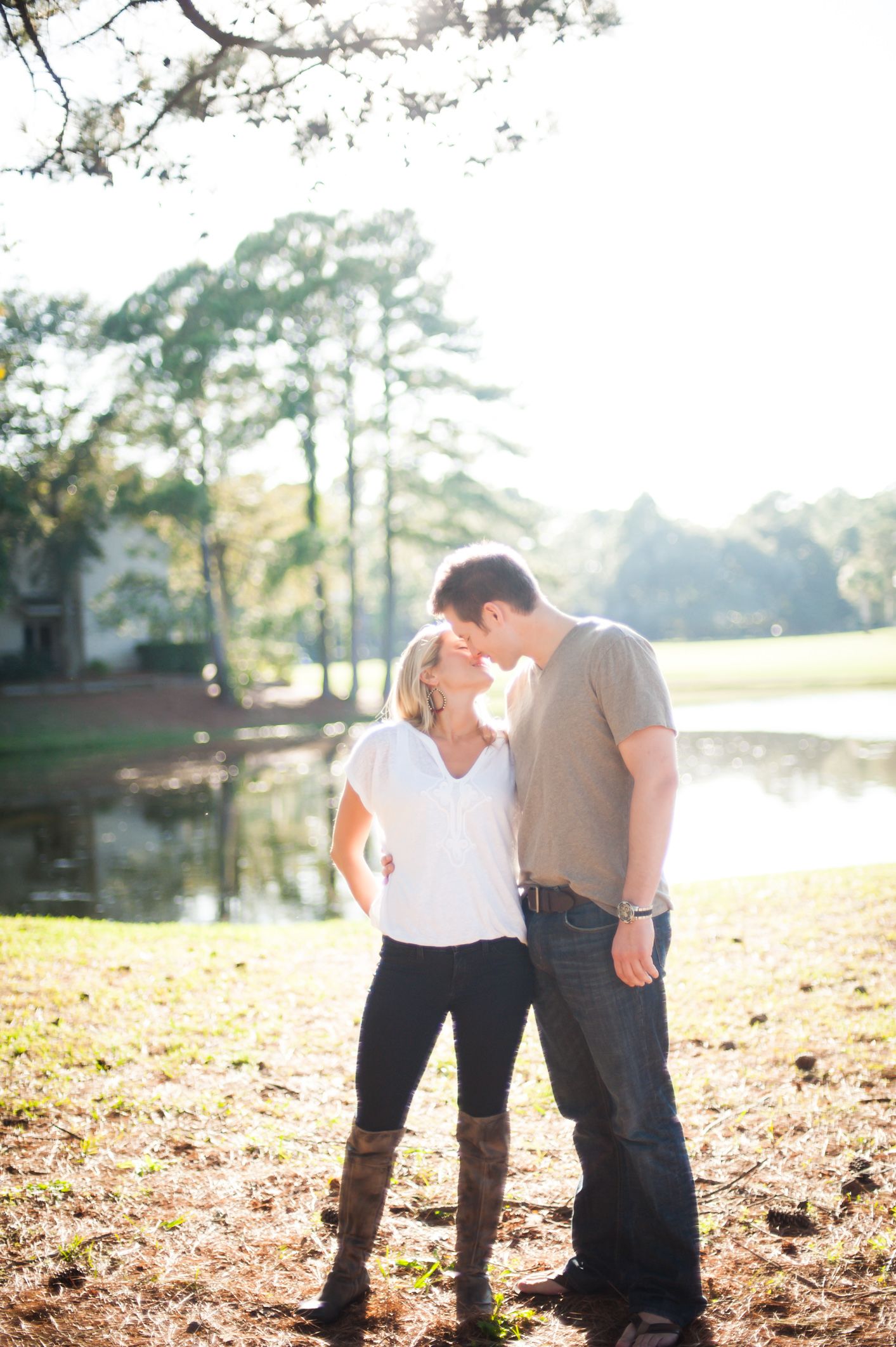 A couple stands together in a sunlit park with autumn leaves scattered on the ground.