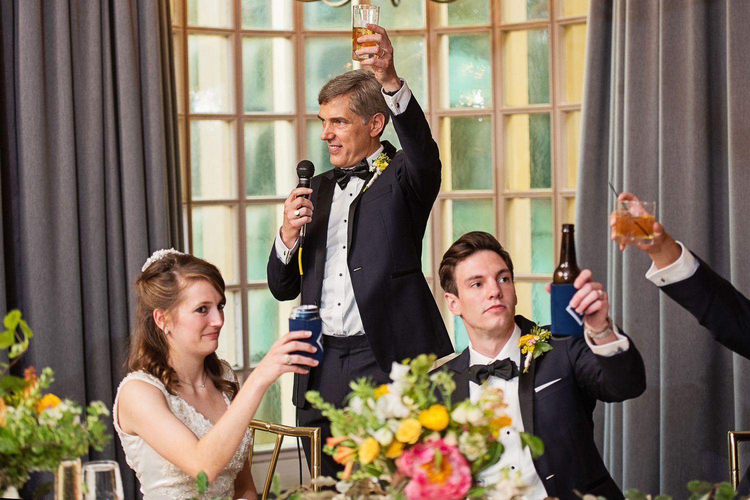 Wedding guests raise their glasses in celebration during a reception toast with colorful floral centerpieces on the table.