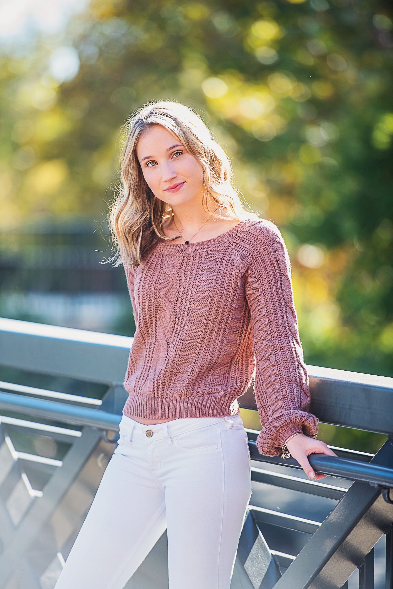 Person in mauve sweater and white pants posing outdoors against autumn foliage.