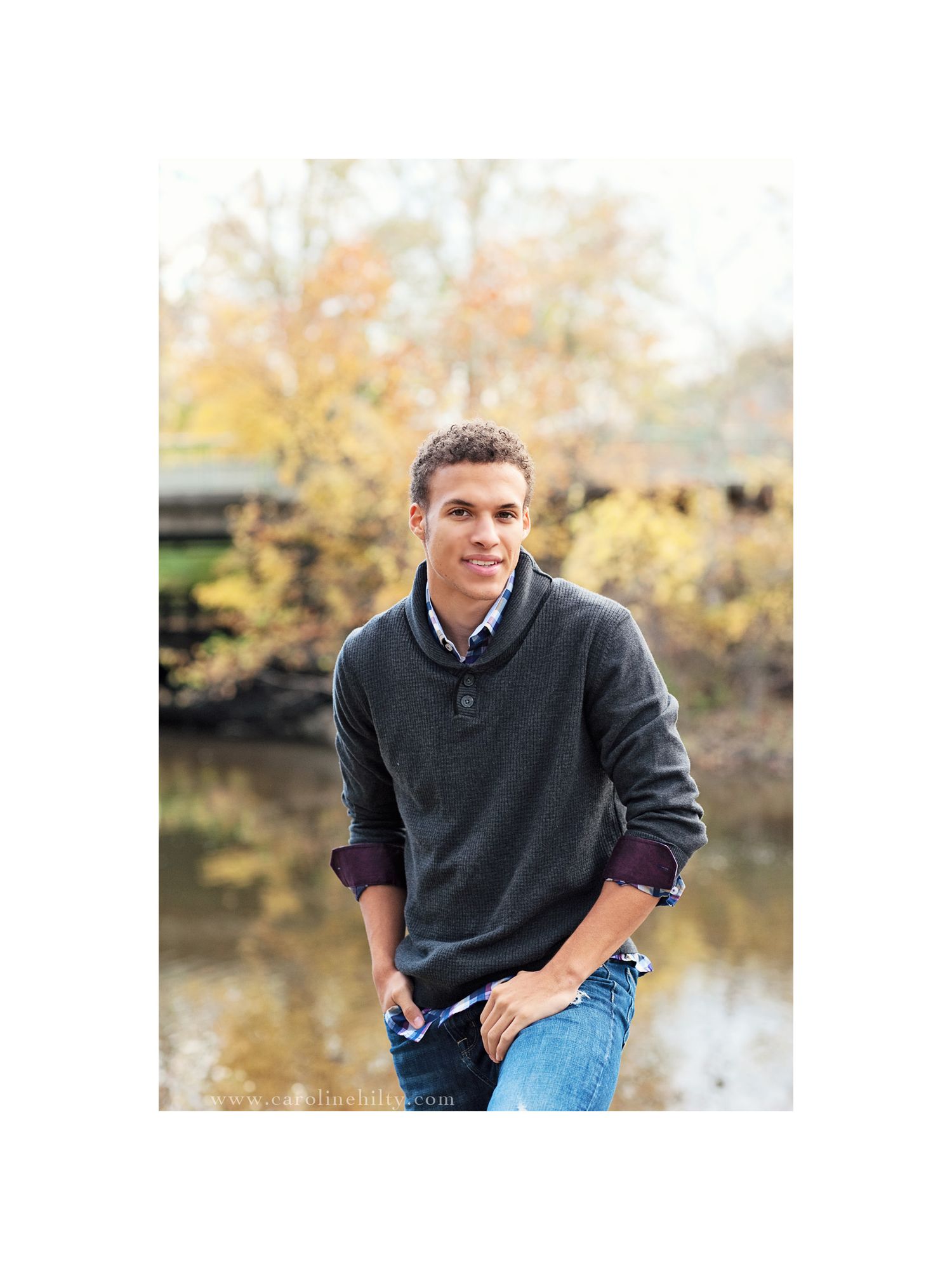 Person in dark sweater and jeans sits casually outdoors during autumn with water and fall foliage in background.