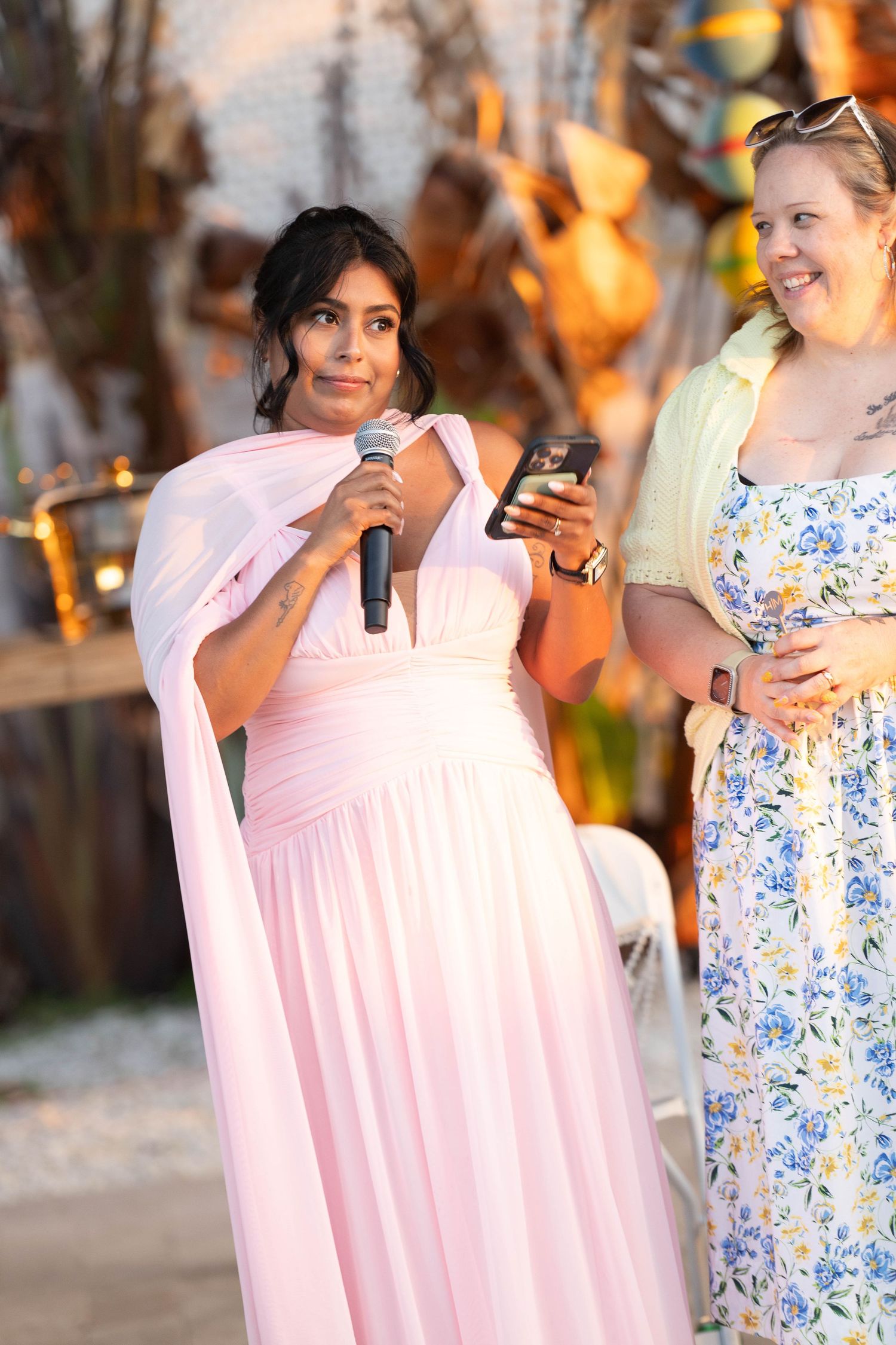 Woman in pink gown speaks into microphone at outdoor event beside smiling woman in floral dress.
