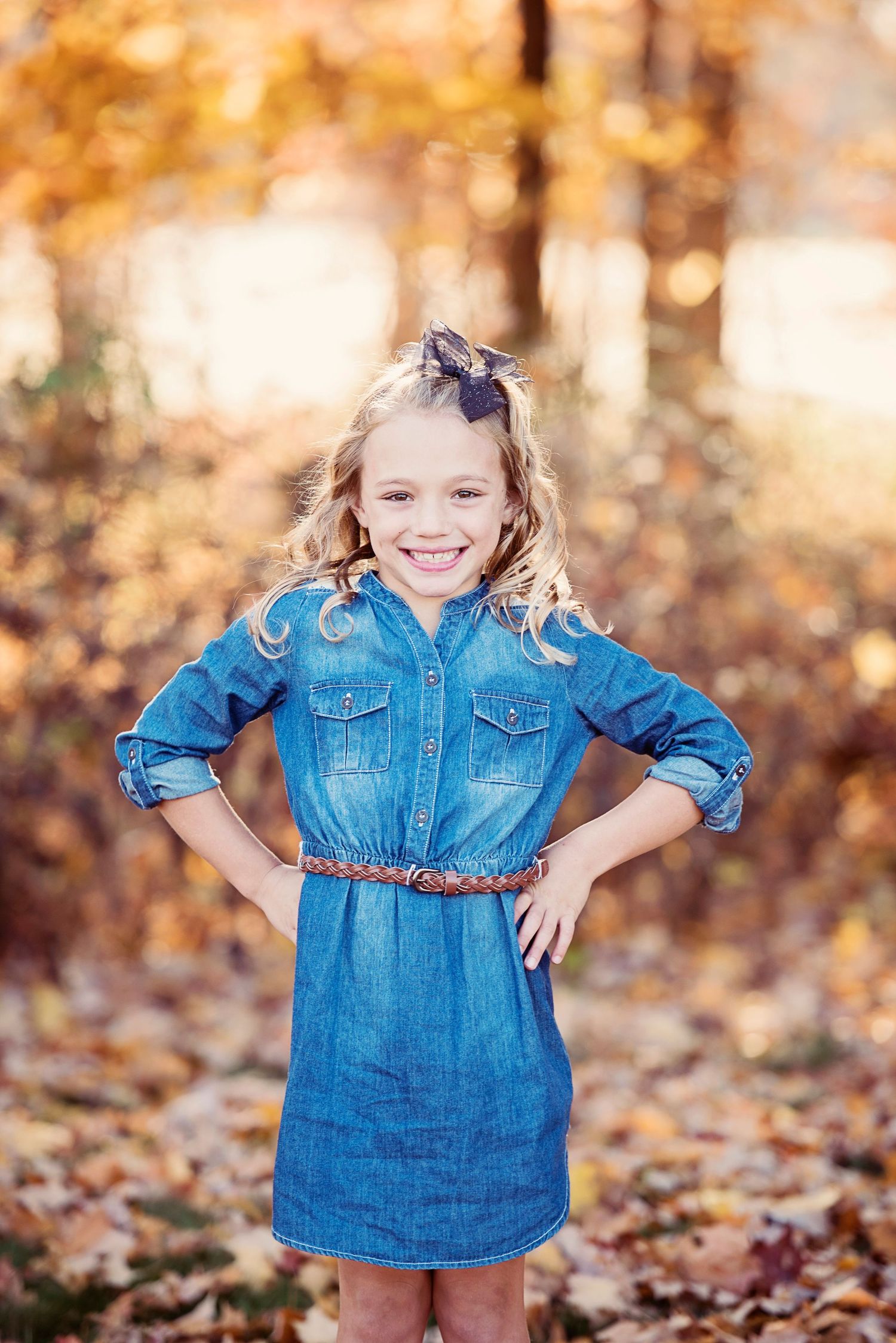 A child wearing a denim dress twirls and plays in autumn leaves during a golden sunset photo session.