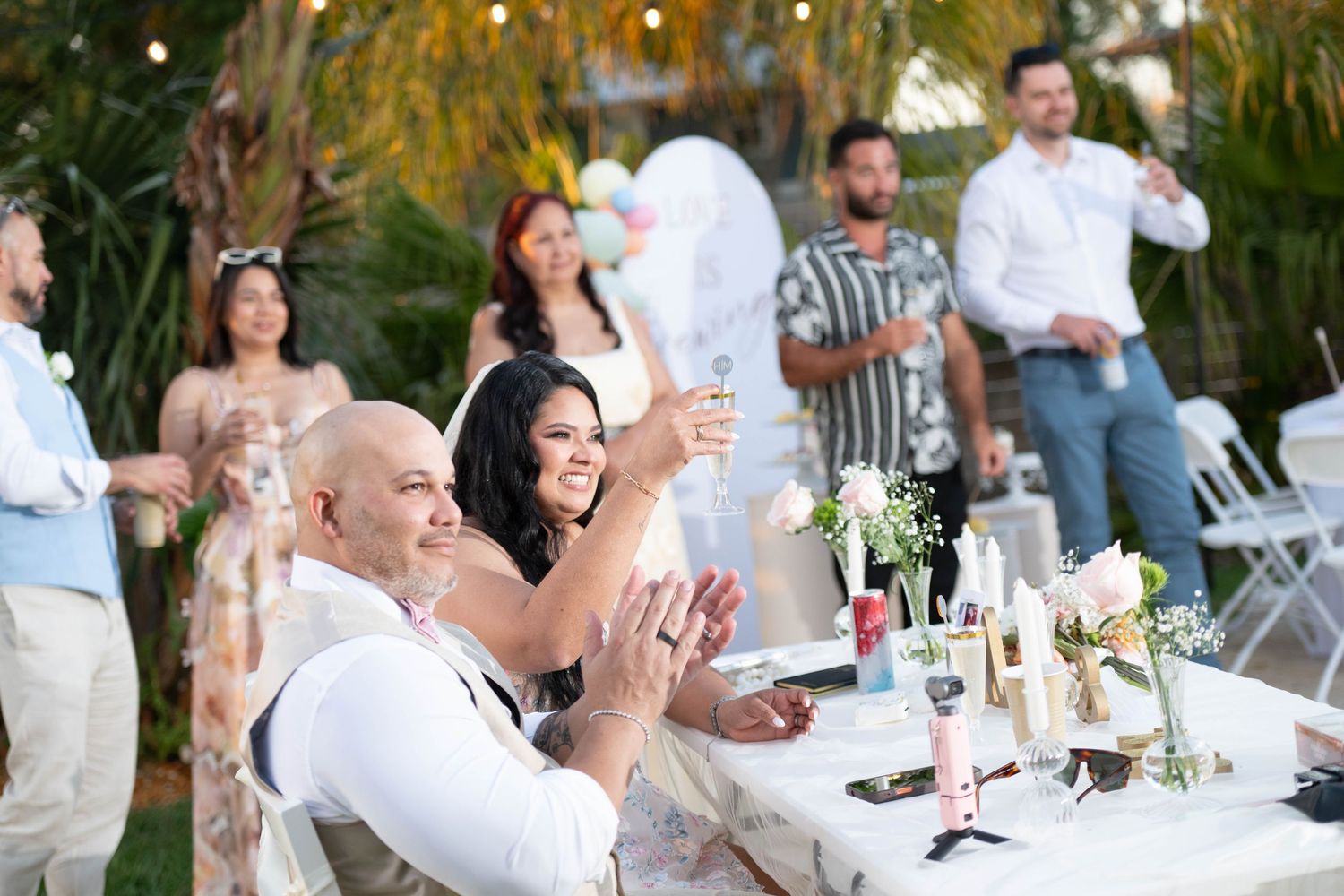 Couple smiling and clapping at outdoor wedding reception table with guests raising glasses in background.