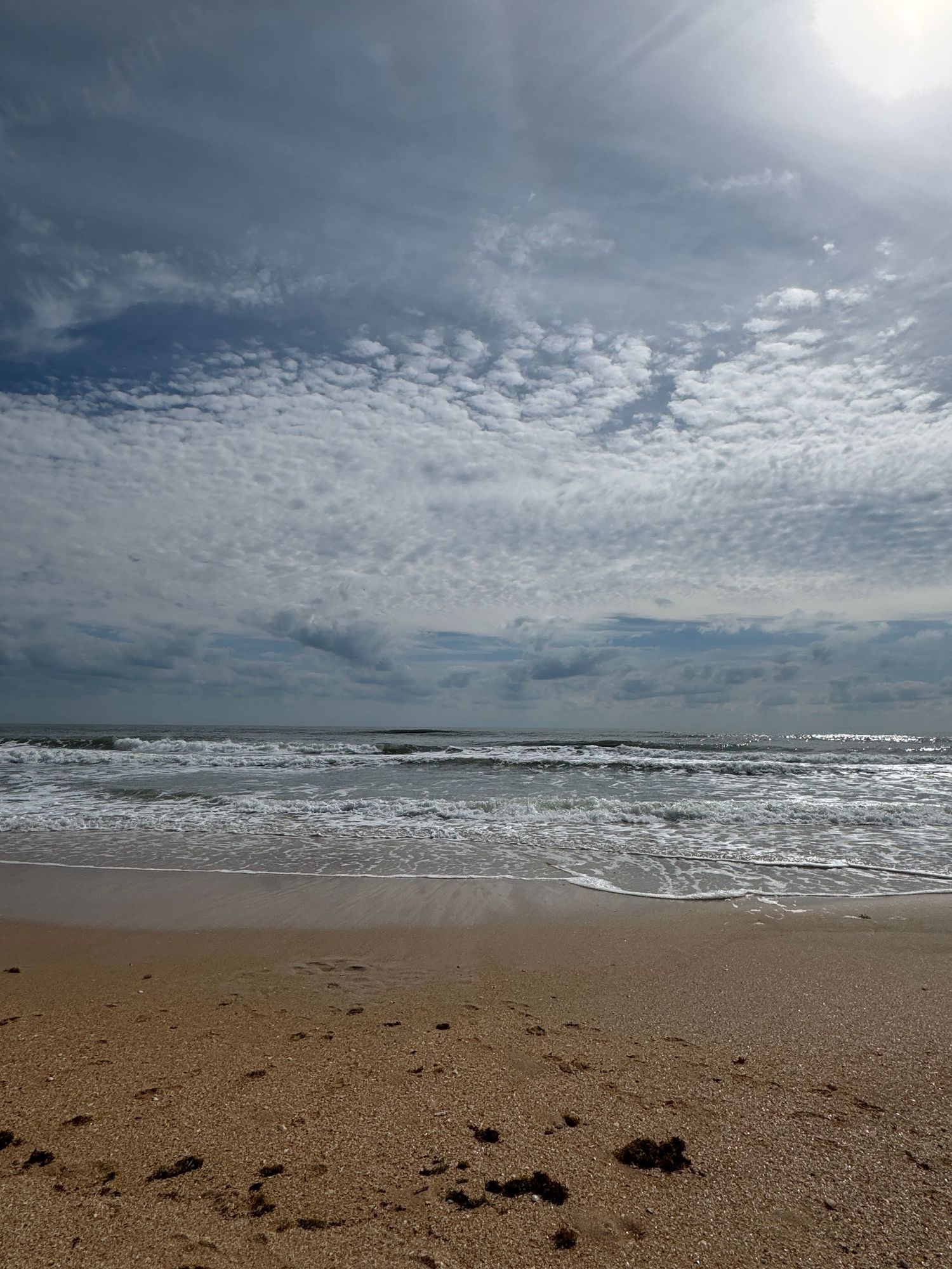 Sandy beach with gentle waves under a dramatic cloudy sky with bright sun, showcasing a serene coastal seascape.