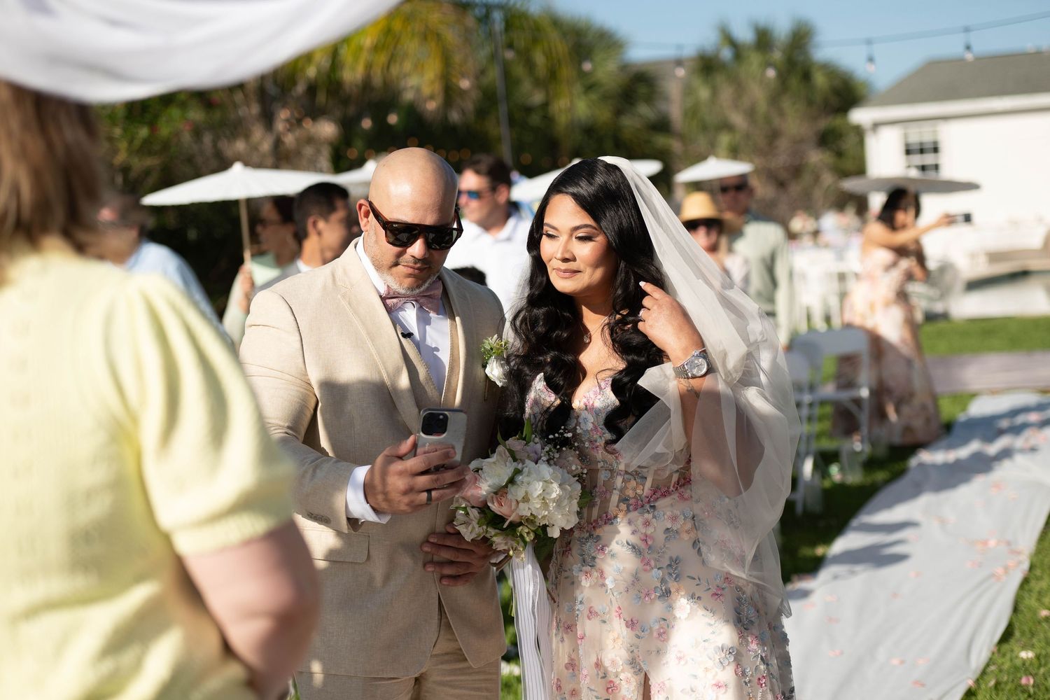 Bride in floral gown and veil walks down outdoor aisle with escort in tan suit at a sunny garden wedding.