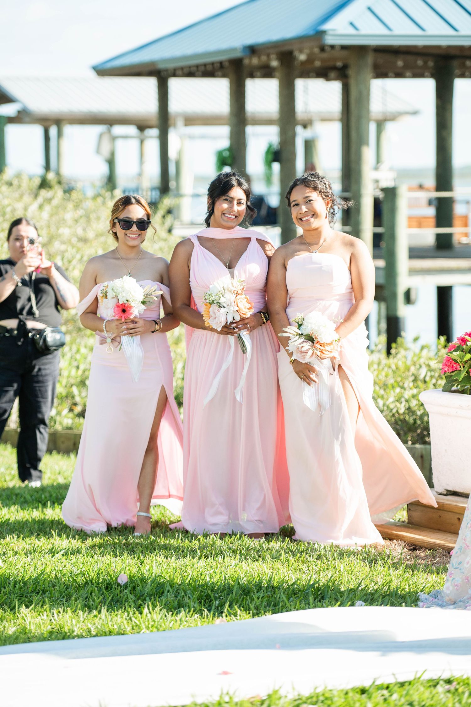 Three bridesmaids in pink gowns holding bouquets smile outdoors at a waterfront wedding ceremony.