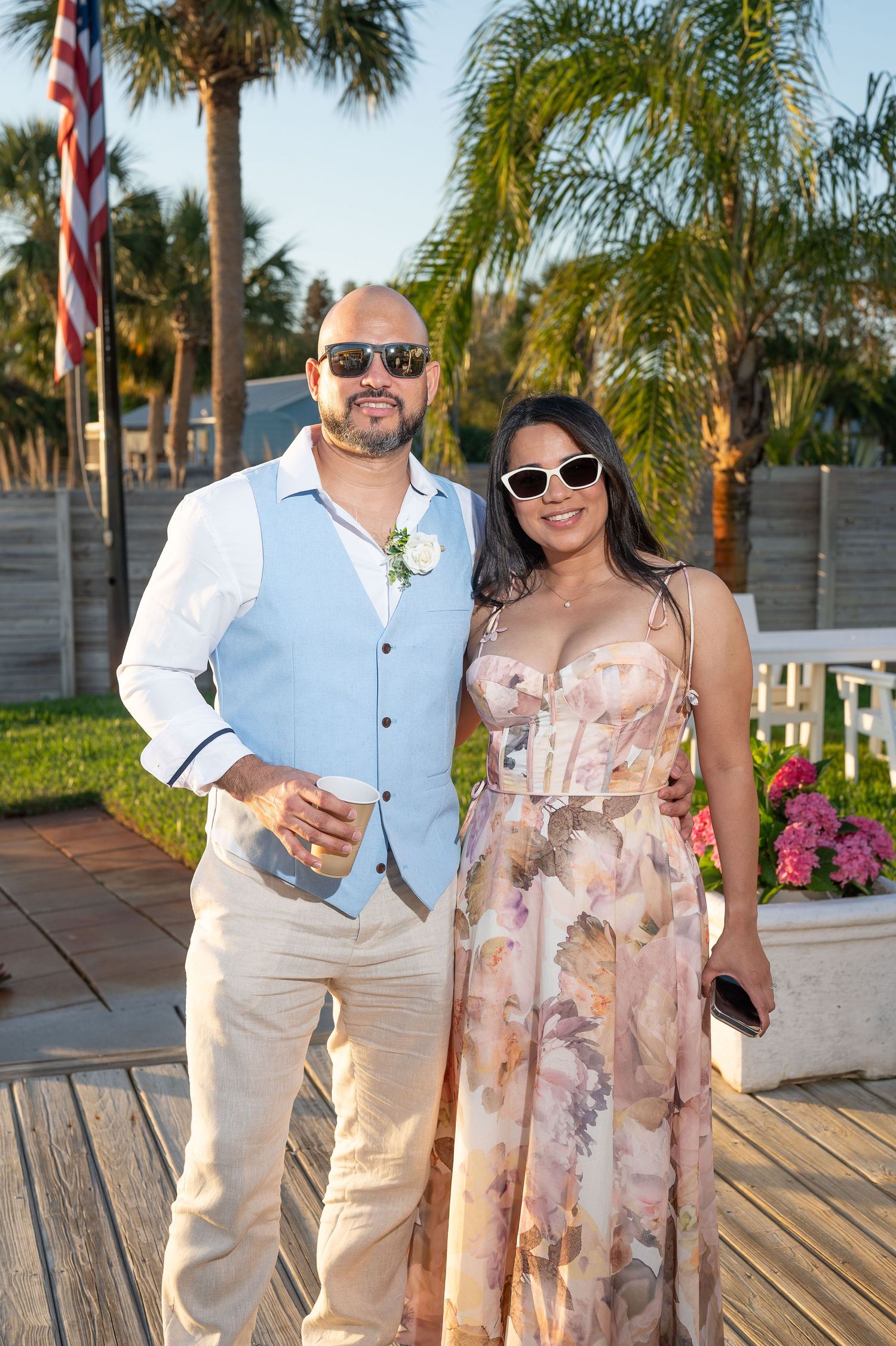 Couple posing outdoors at a tropical wedding venue, man in blue vest and woman in floral maxi dress.