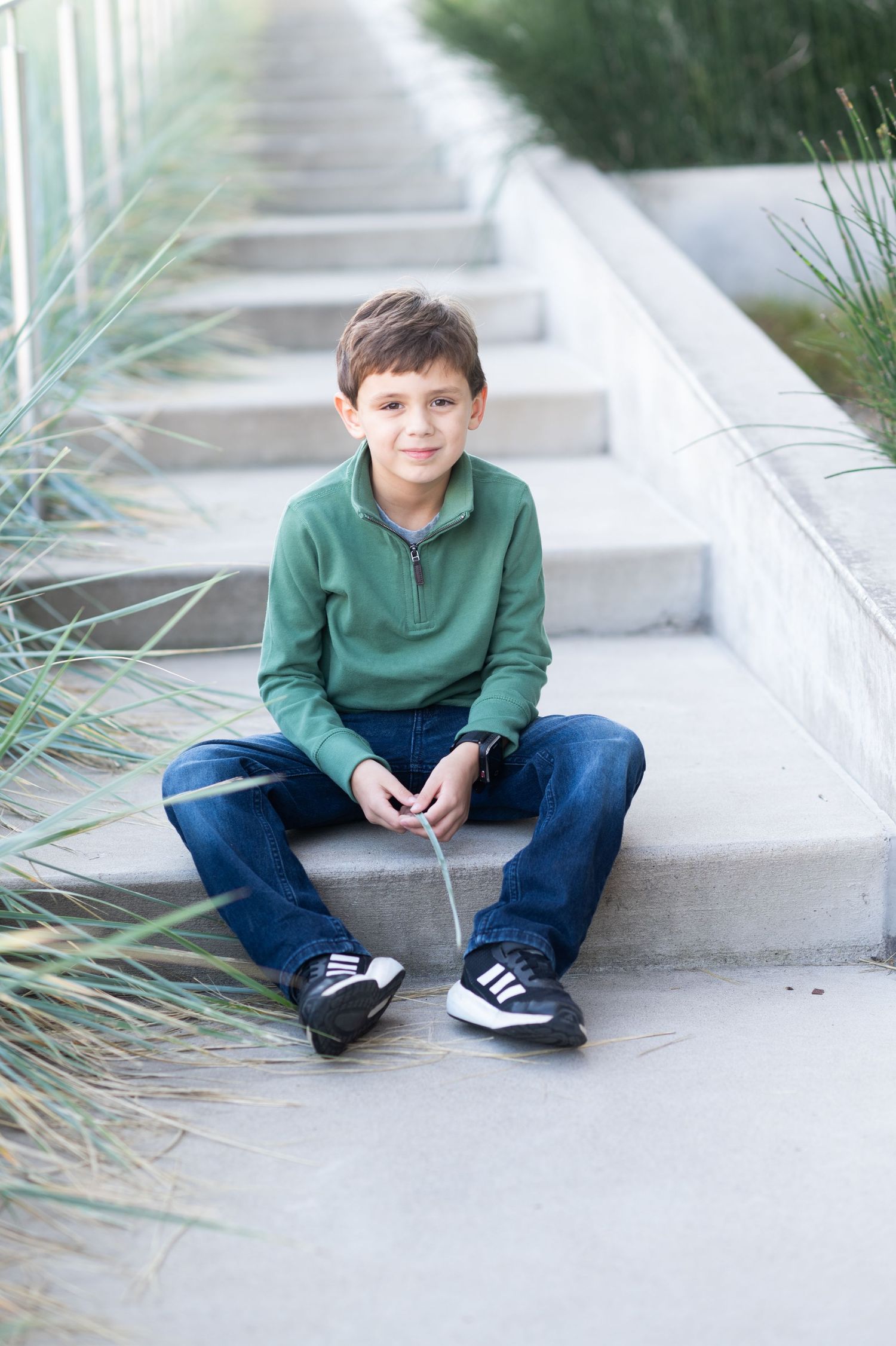 A young boy in a green sweatshirt and jeans sits cross-legged on concrete steps outdoors, smiling at the camera.