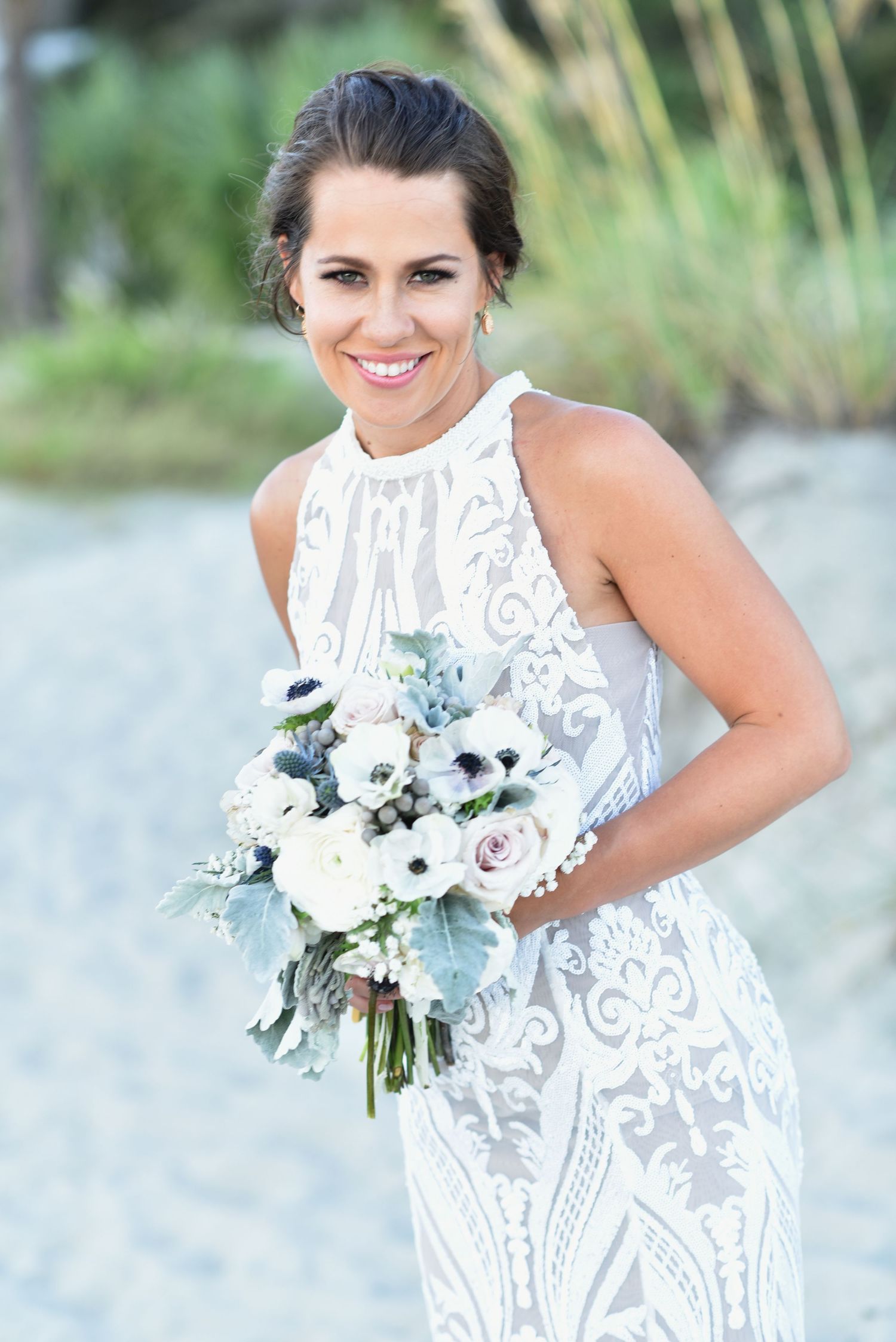 Smiling bride in white lace halter gown holding elegant bouquet of white anemones and roses outdoors.