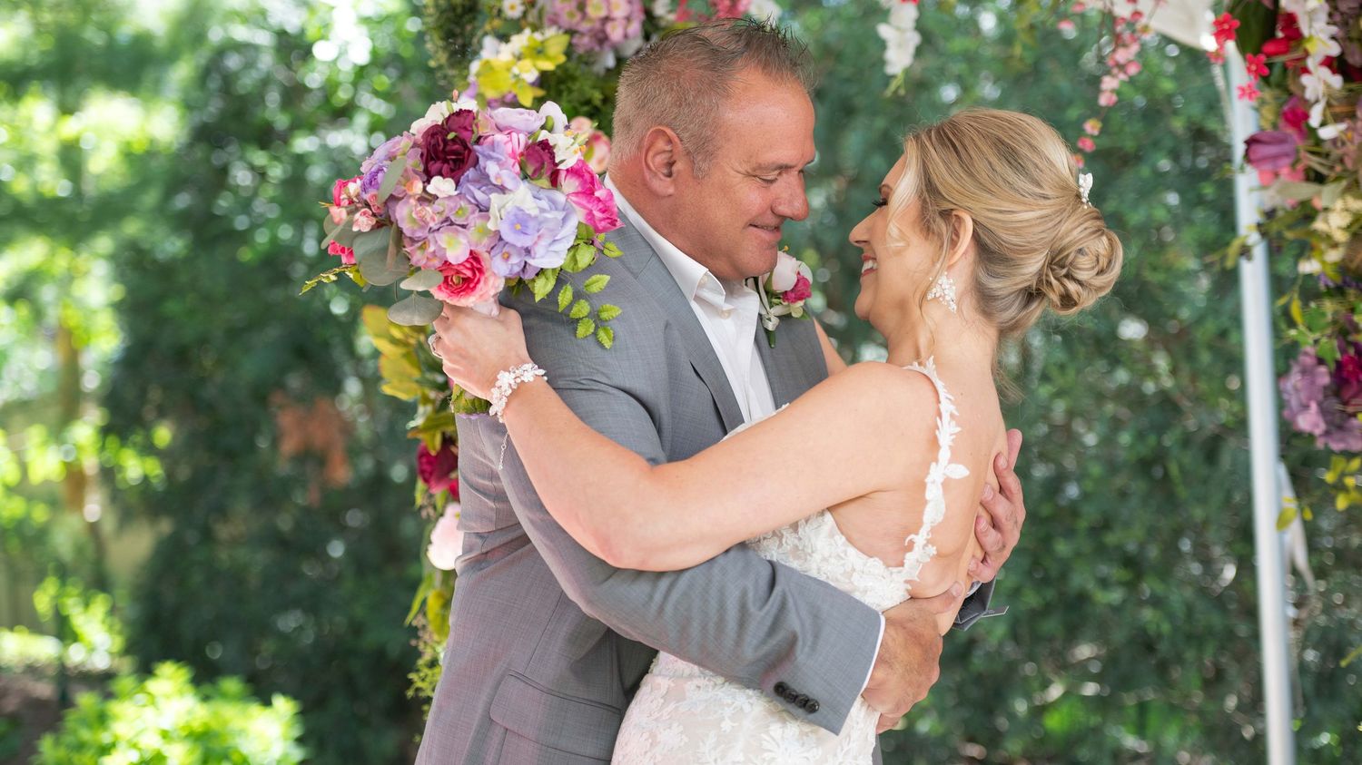 A bride and groom share a romantic embrace outdoors under a floral arch during their wedding ceremony.
