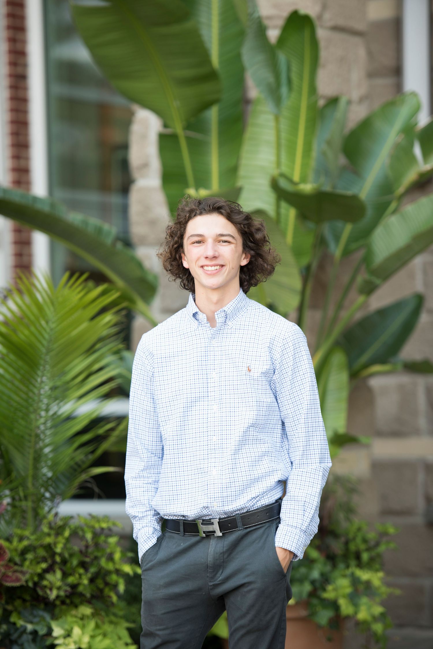A person in a blue checkered shirt and dark pants poses outdoors near tropical plants and a brick building.