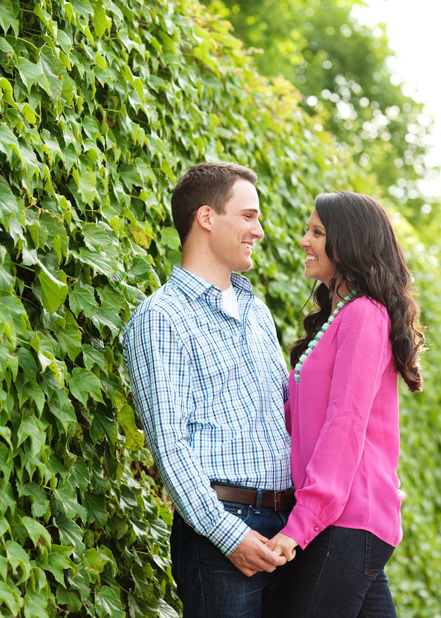 A couple shares a romantic moment while holding hands against a lush green ivy wall background.
