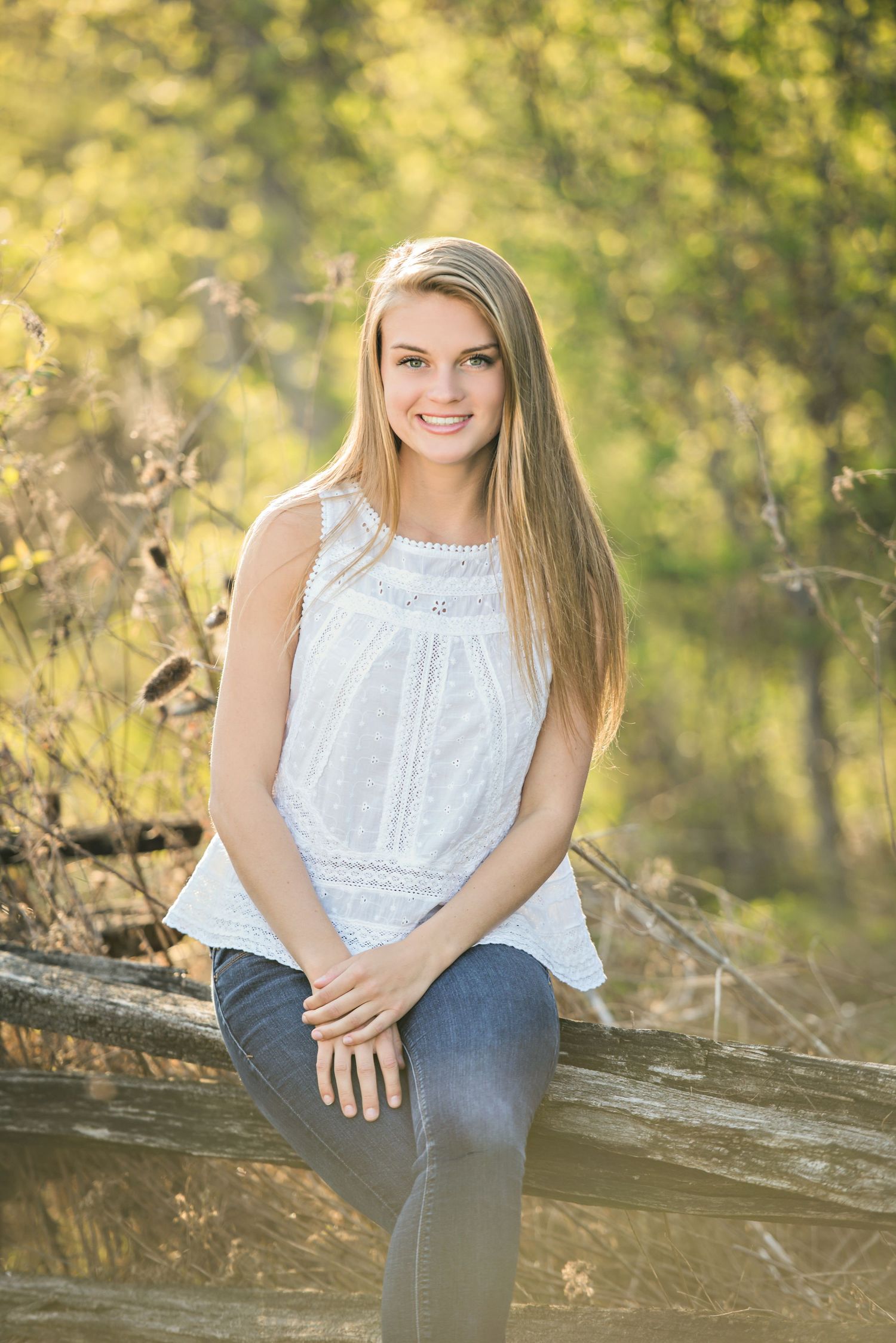 A person in a white sleeveless top and jeans poses outdoors on a fallen log with golden sunlit foliage in the background.
