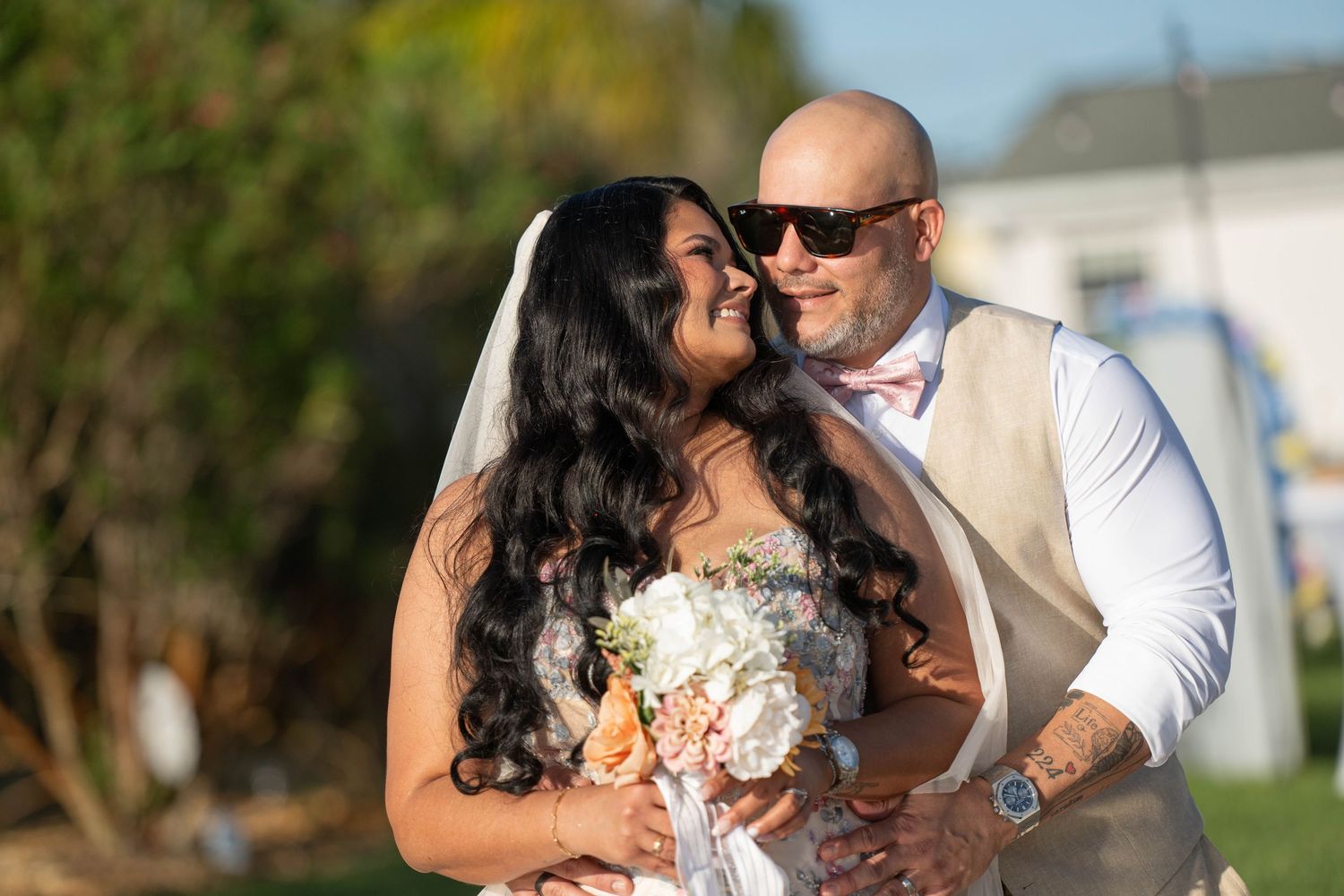 Bride and groom embrace outdoors, bride holding white bouquet wearing veil, groom in vest and sunglasses on wedding day.