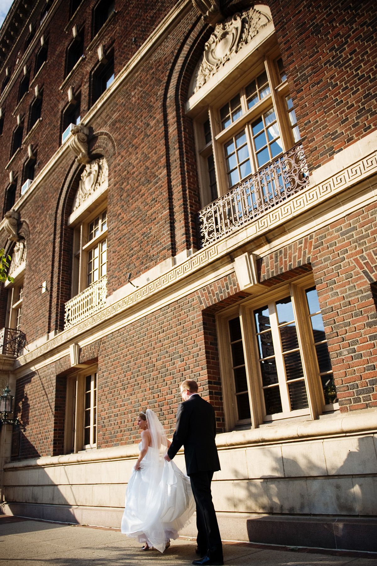 A couple in formal attire walks along a brick building with ornate architectural details and windows.