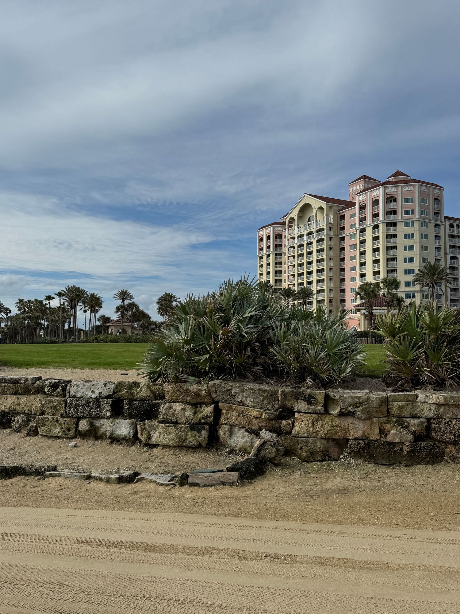 Beachside resort tower with tropical palms, stone retaining wall, and sandy path under cloudy blue skies.