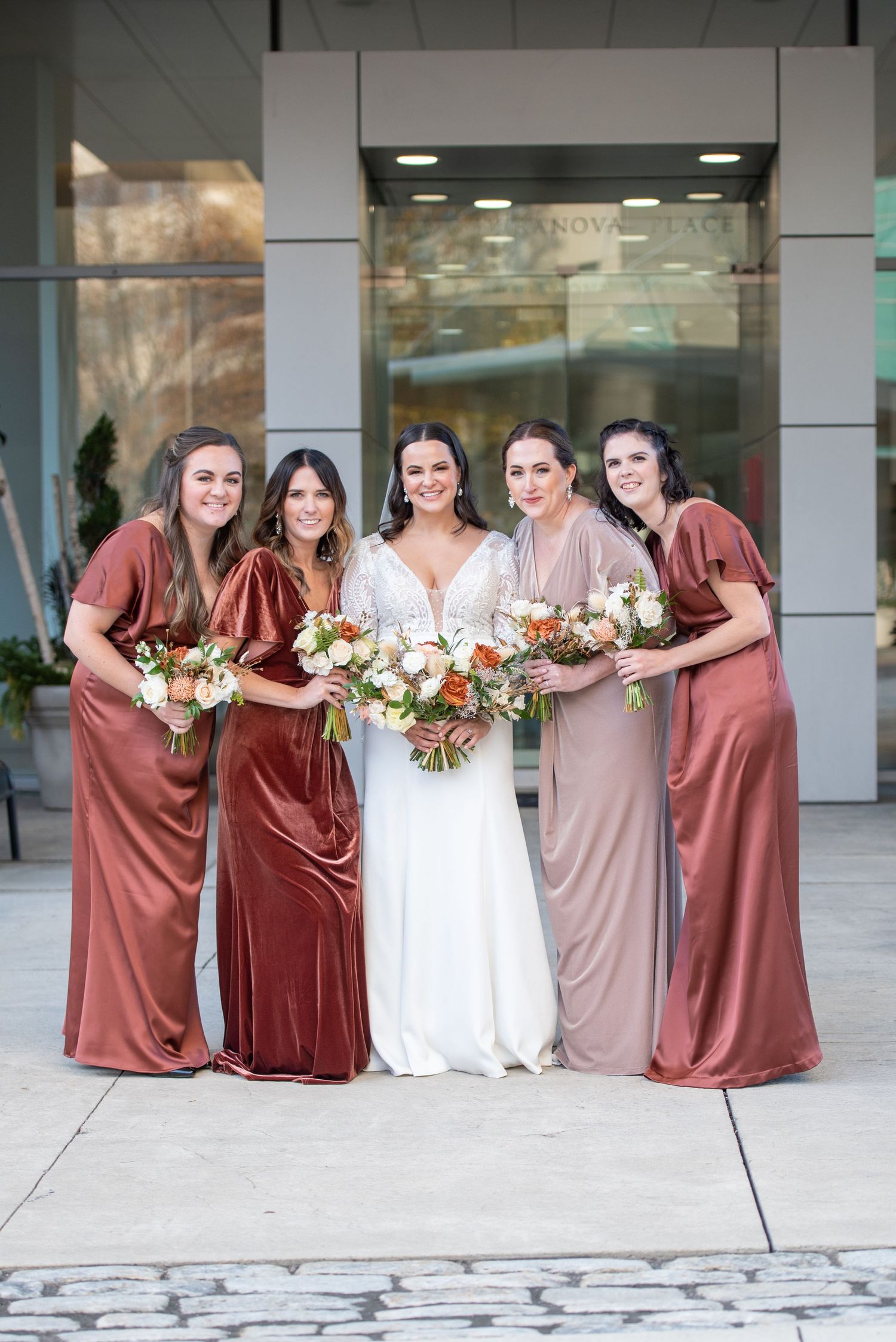Bridal party in flowing velvet dresses in shades of rust and mauve pose together holding floral bouquets.