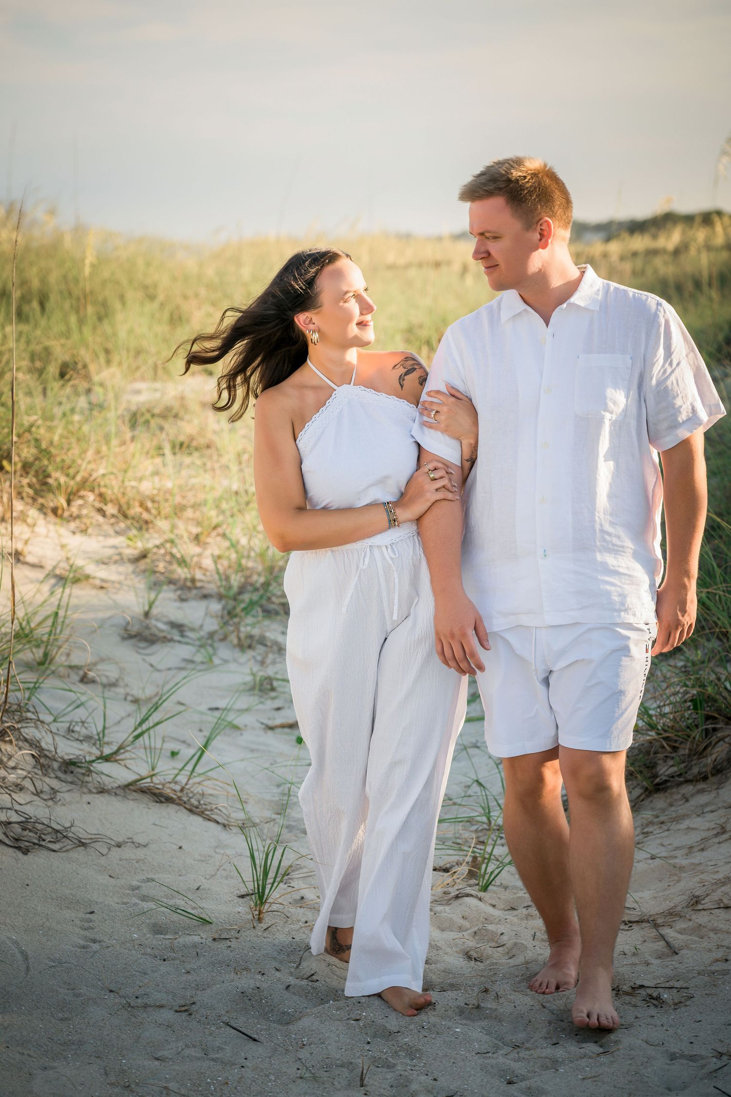 Couple in matching white outfits walking barefoot on sandy beach dunes at golden hour sunset.