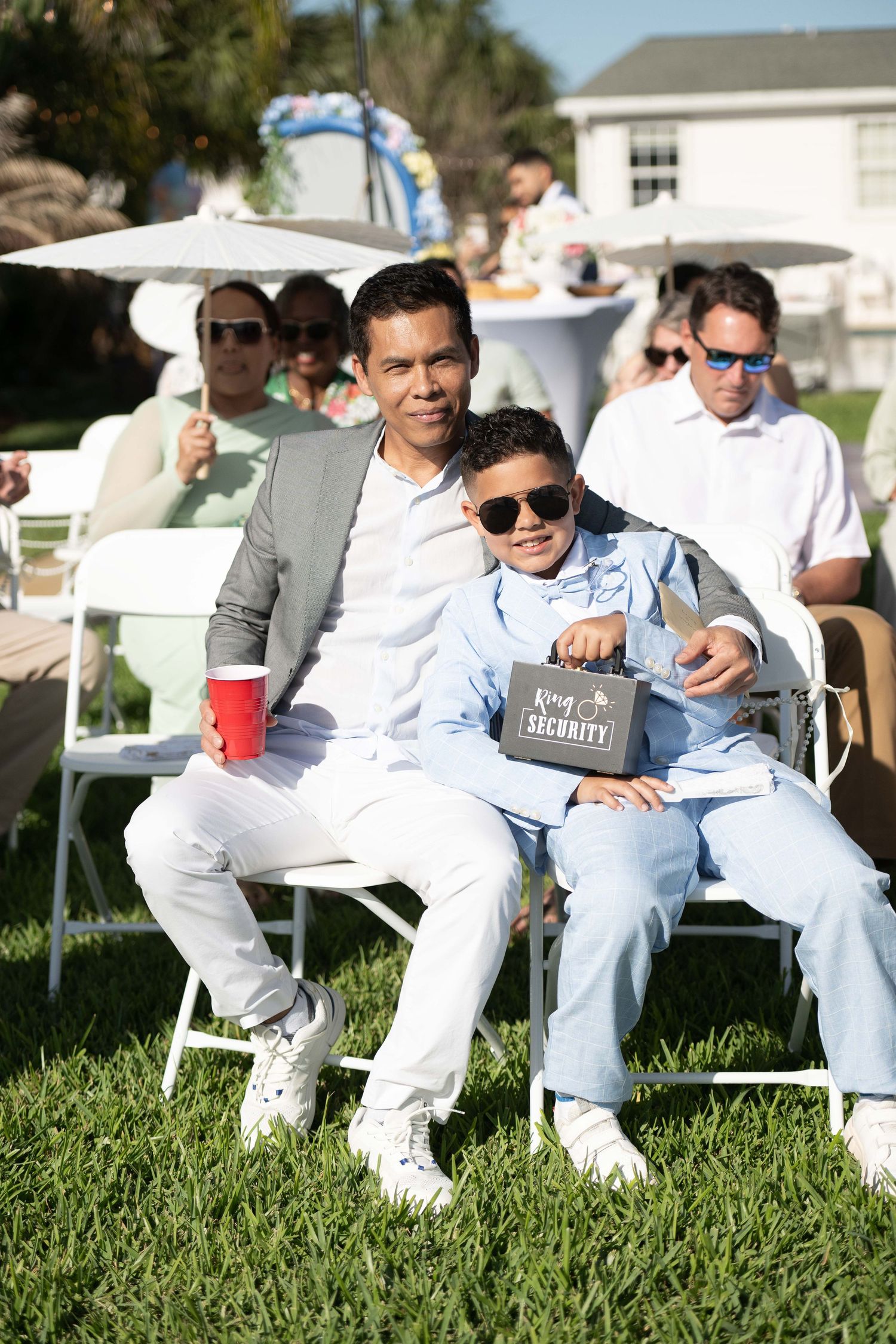A man and boy in sunglasses pose at an outdoor event, boy holds a Ring Security sign, guests seated on white chairs behind them.