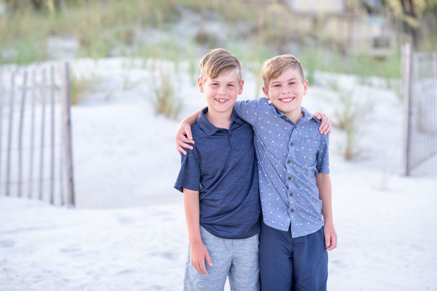 Two smiling boys pose together on a sandy beach, one with arm around the other, wearing casual shirts on a sunny day.