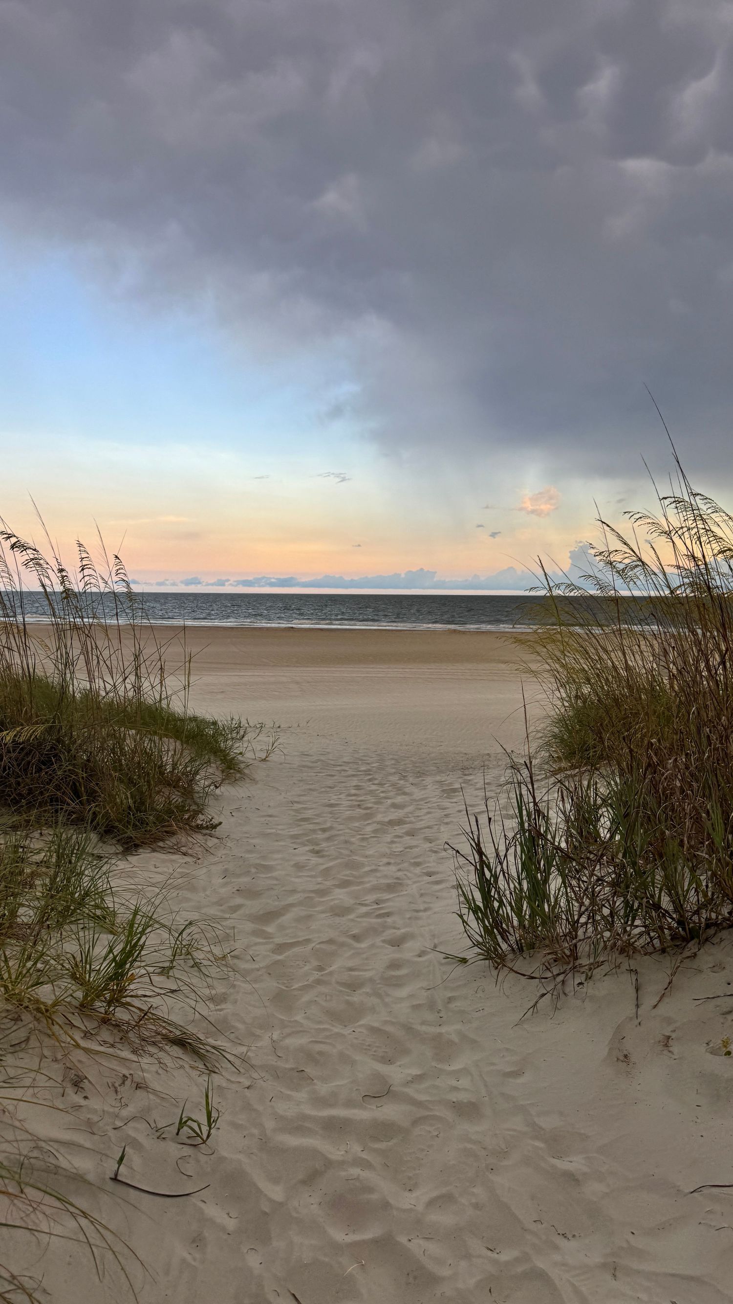 Sandy beach path flanked by sea oats leads to calm ocean shore under dramatic cloudy sunset sky.