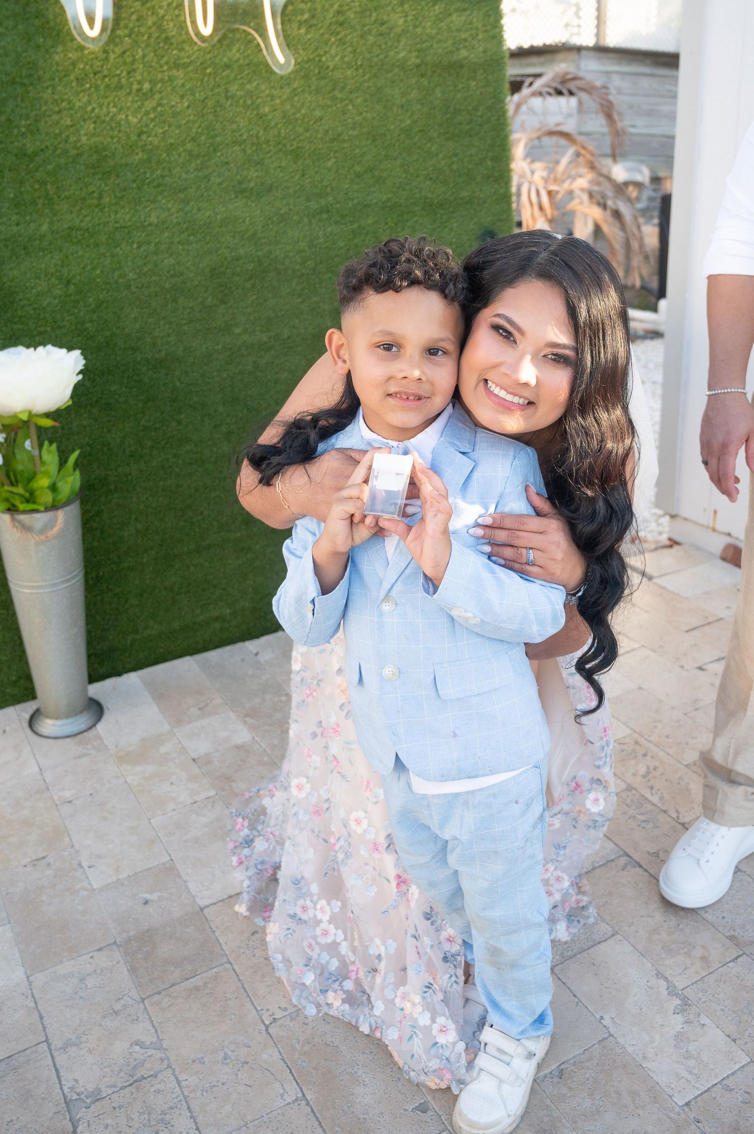 A woman in a floral dress hugs a young boy in a light blue suit at an outdoor event.