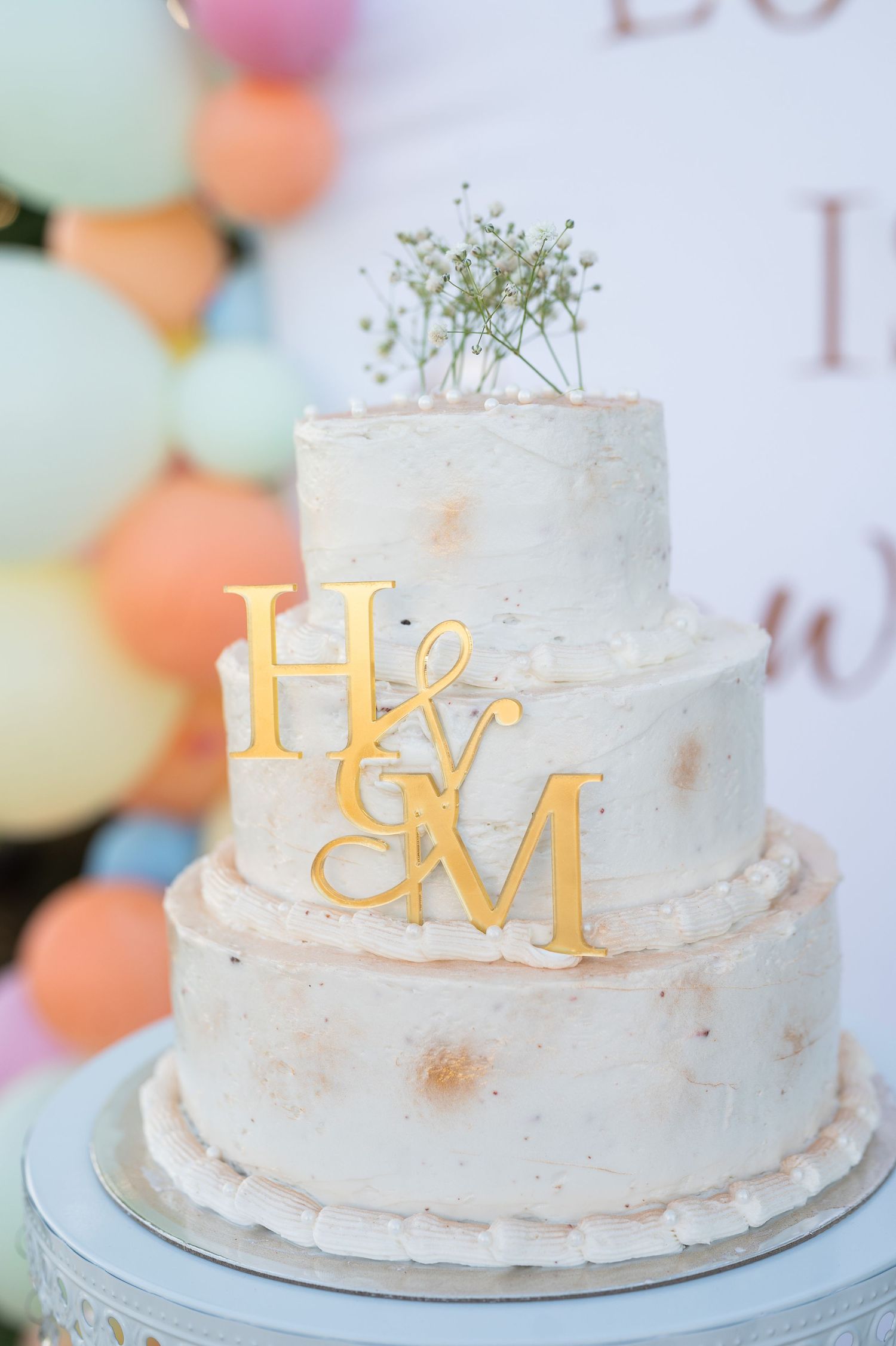 Two-tier white wedding cake with gold H&M monogram topper and baby's breath, displayed against colorful balloon backdrop.