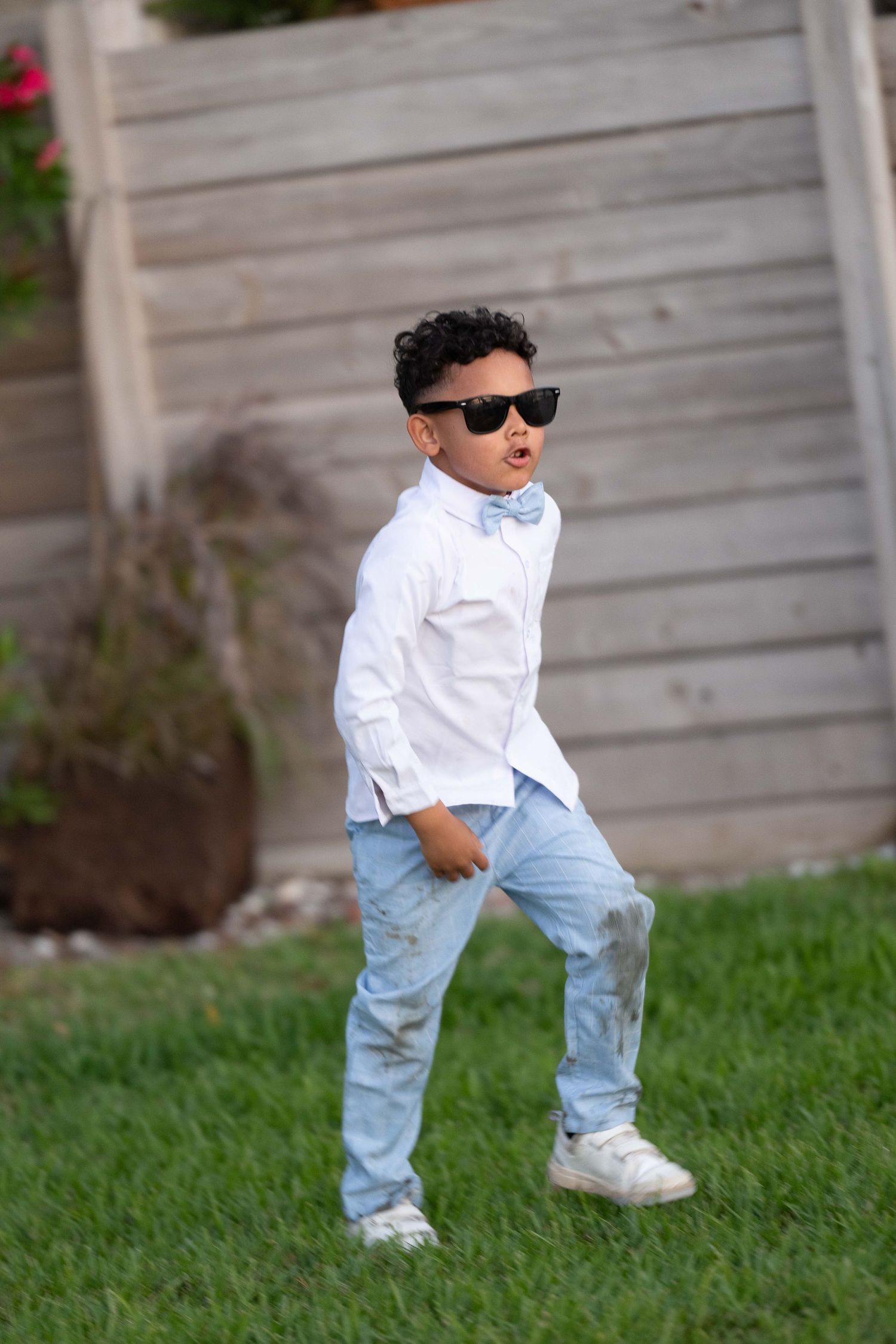 Stylish young boy in white shirt, bow tie, jeans, and sunglasses posing confidently on green grass outdoors.