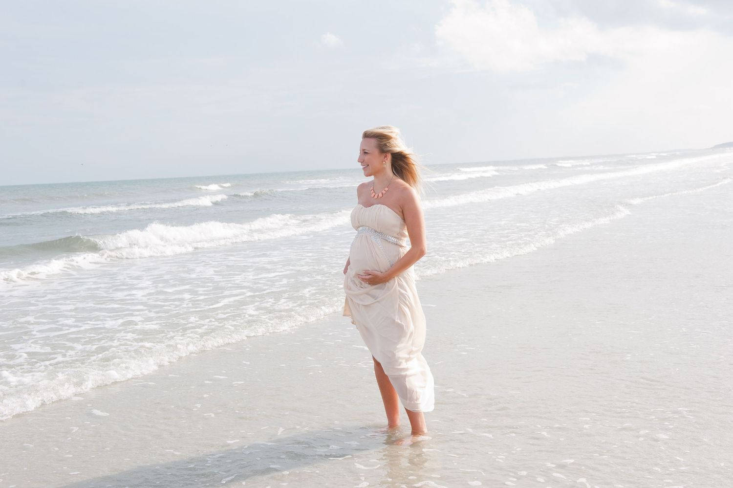 Pregnant woman in white dress standing on a sunny beach, cradling her baby bump while ocean waves wash over her feet.