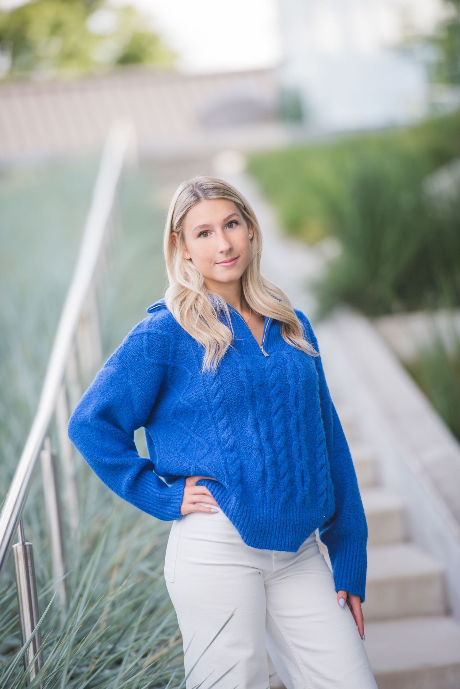 Blonde woman in blue cable-knit sweater and white pants posing on outdoor steps with greenery.