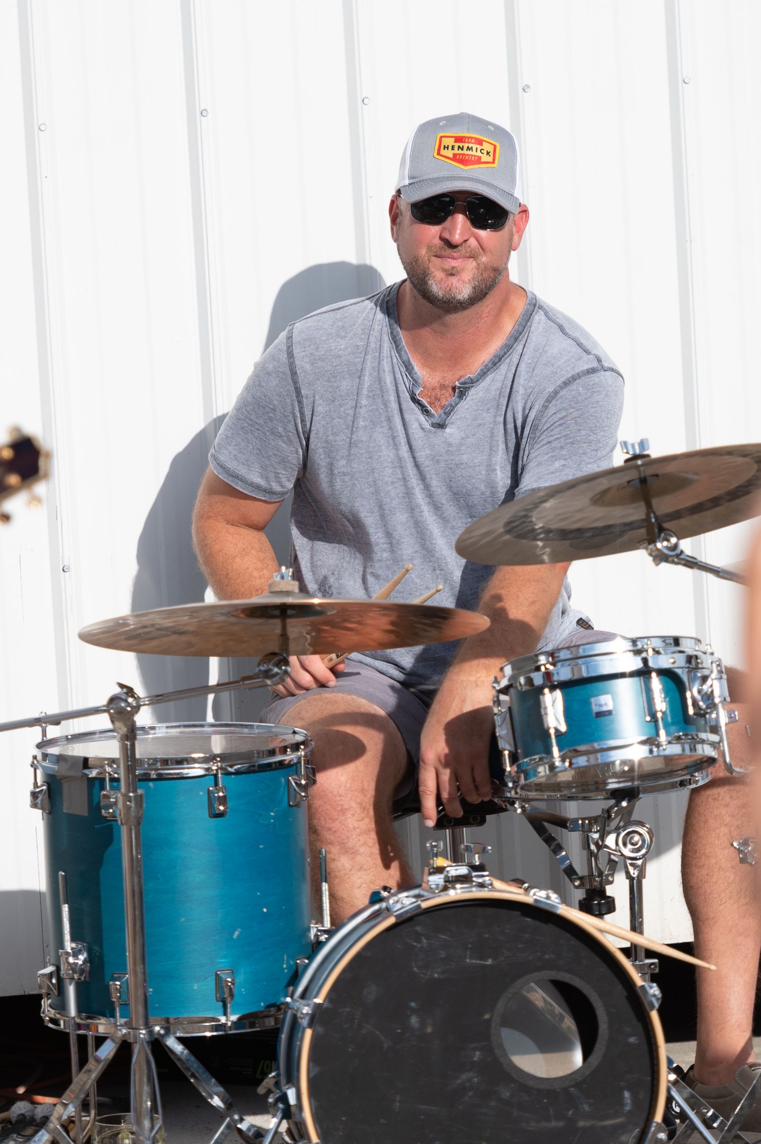 Drummer in gray t-shirt and sunglasses playing on a blue drum kit against a white backdrop.