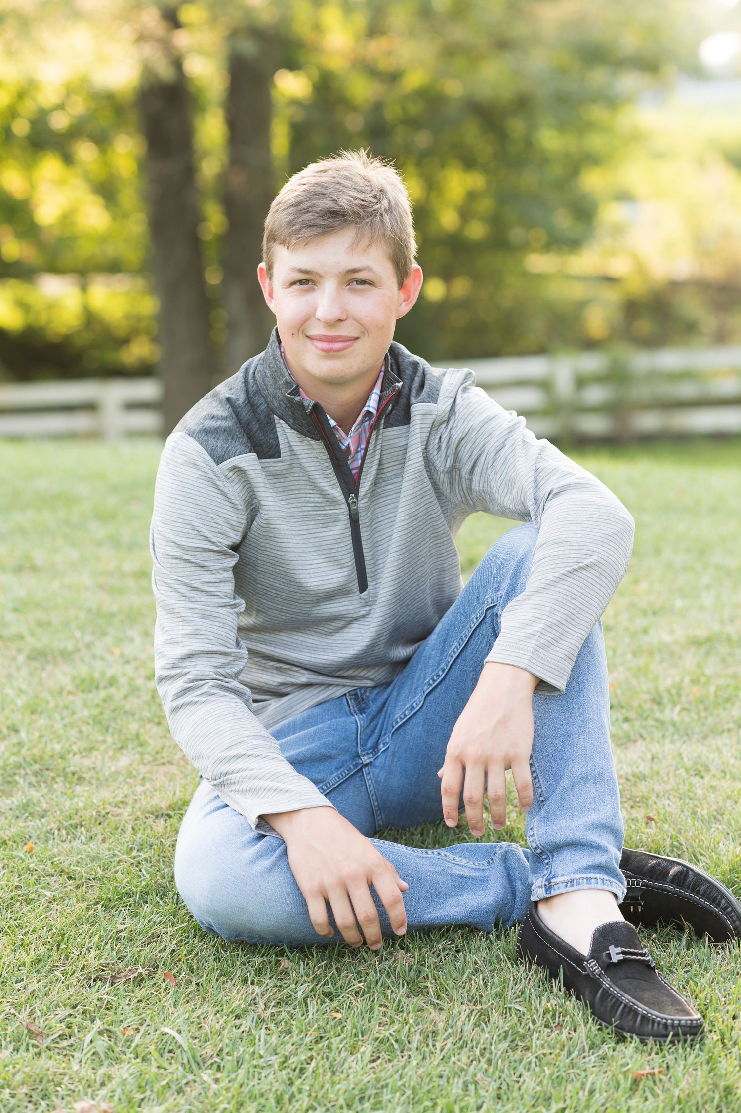 A person in a gray pullover and jeans sits casually on green grass during a sunny outdoor photo session.