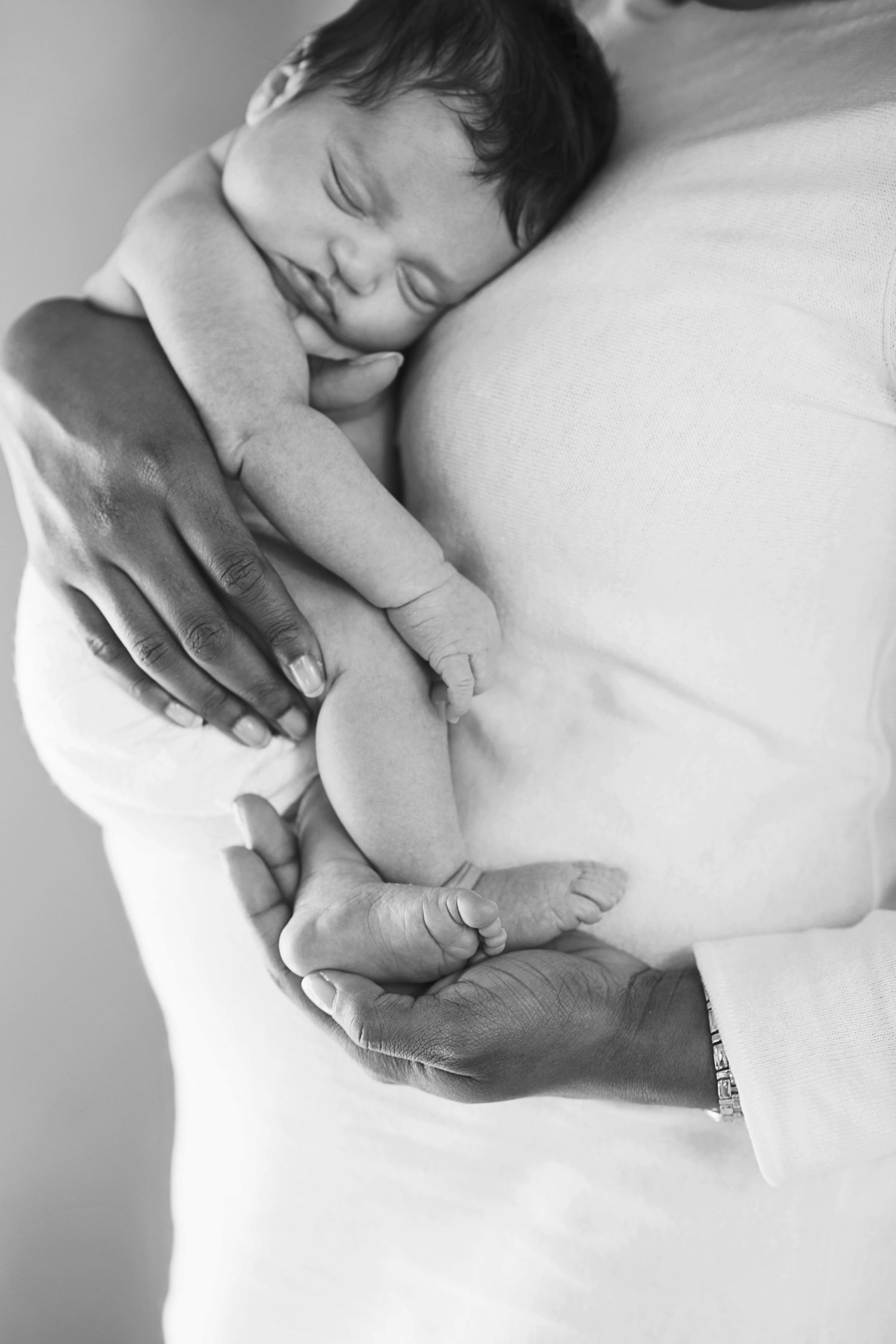 Black and white close-up shots of gentle hands cradling a sleeping newborn baby on white bedding.