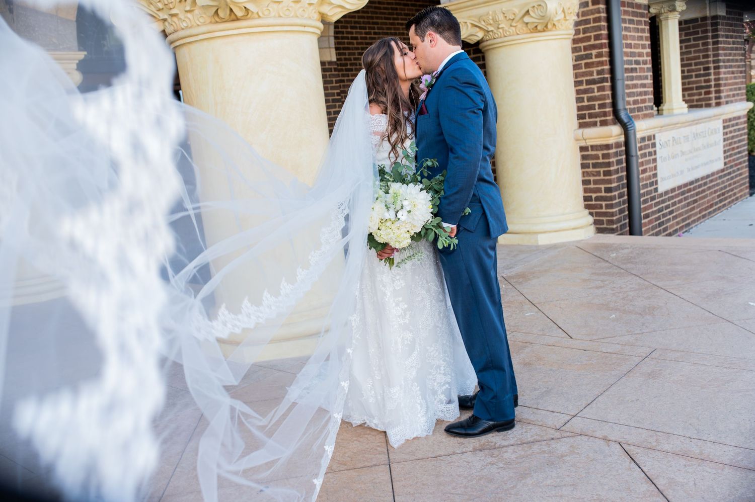 A bride in a flowing white veil and groom in navy suit embrace in front of a brick building.